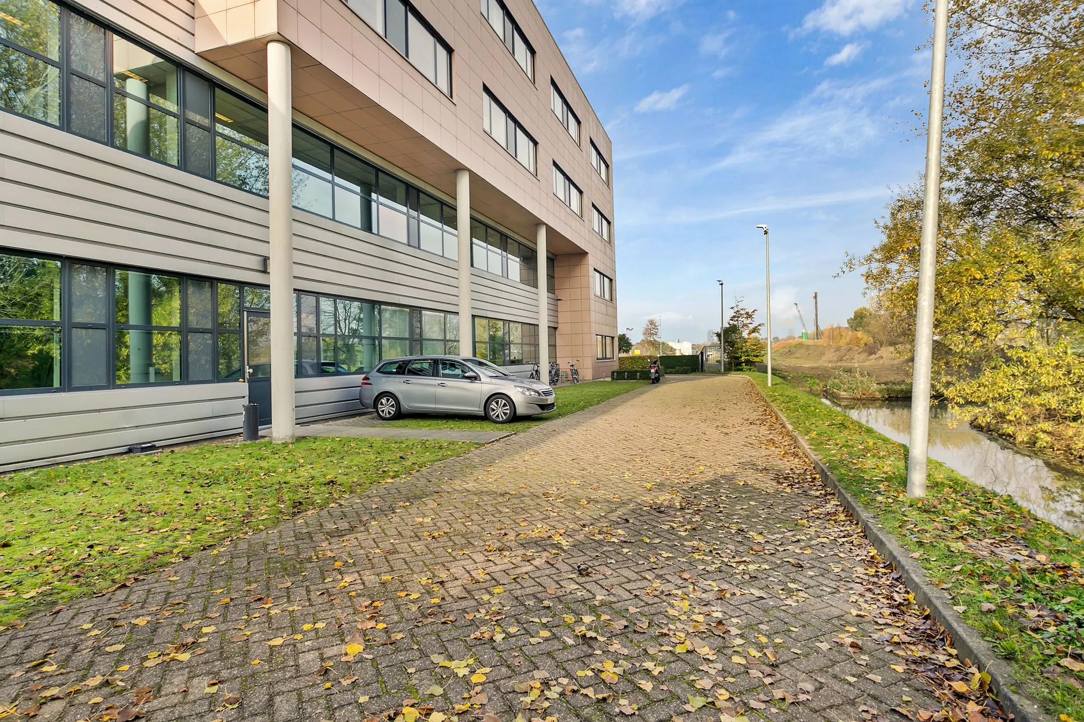 Kantoorgebouw aan de Laan van ’s-Gravenmade met geparkeerde auto en naastgelegen watergang op een herfstdag.