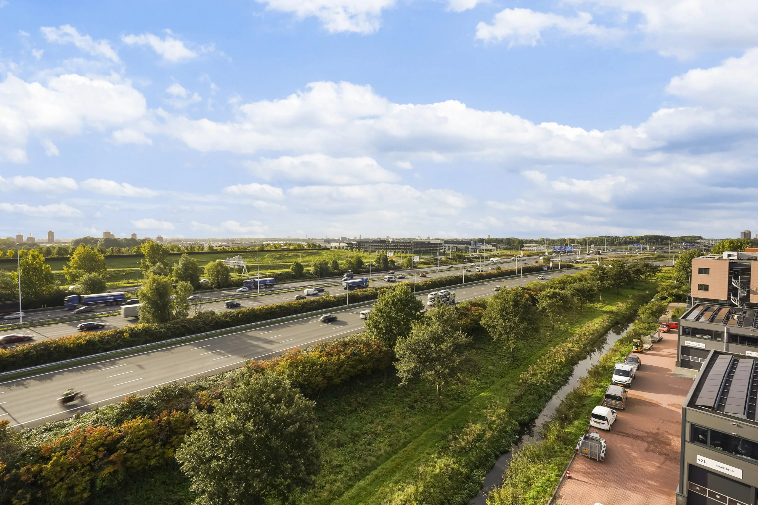 Drukke snelweg gezien vanaf de Laan van 's-Gravenmade met naastgelegen bedrijven en groenstroken onder een deels bewolkte lucht.