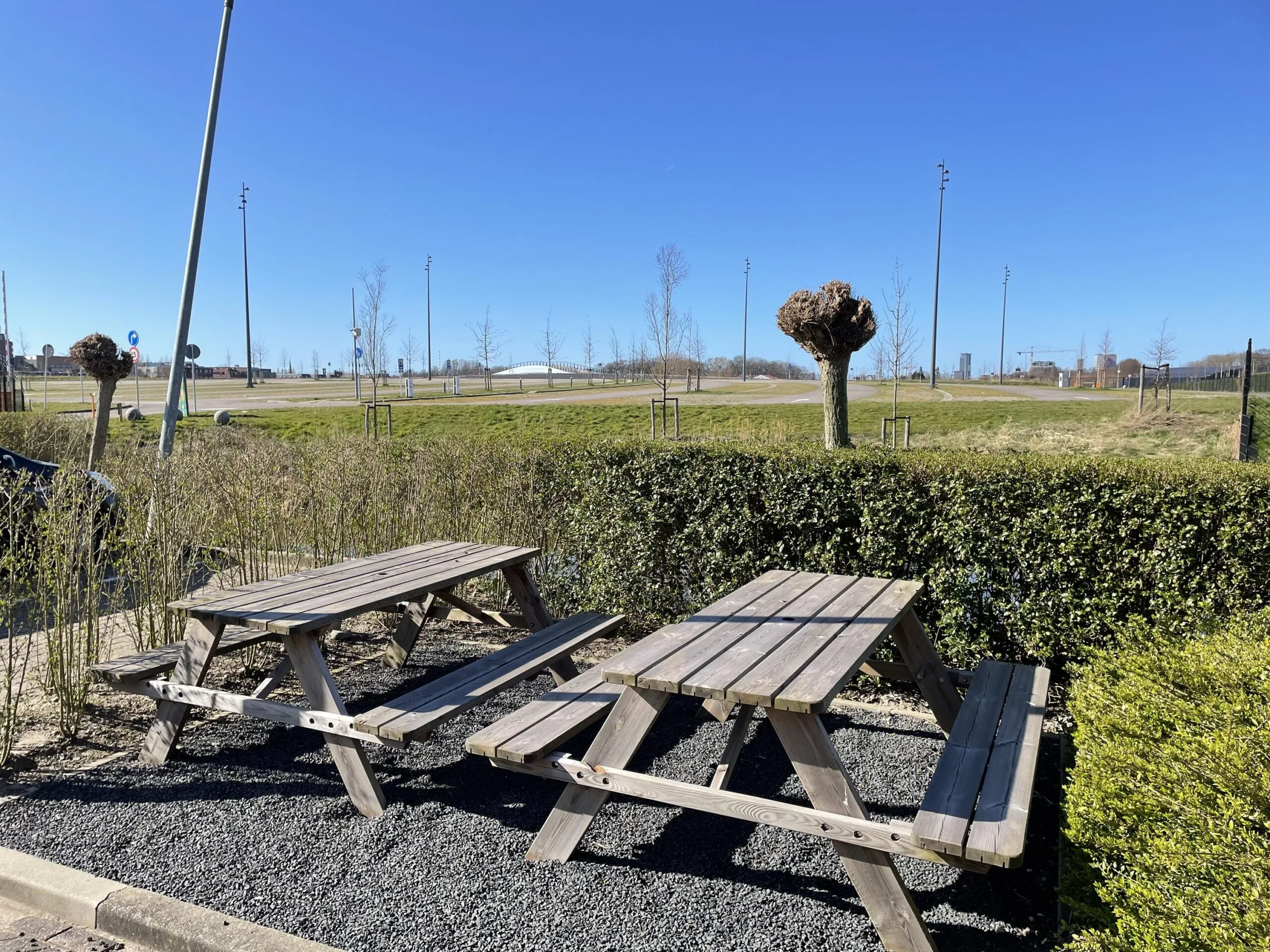 Twee picknicktafels staan op een grindterras aan de Laan van 's-Gravenmade met uitzicht op een open groen veld en een heldere blauwe lucht.