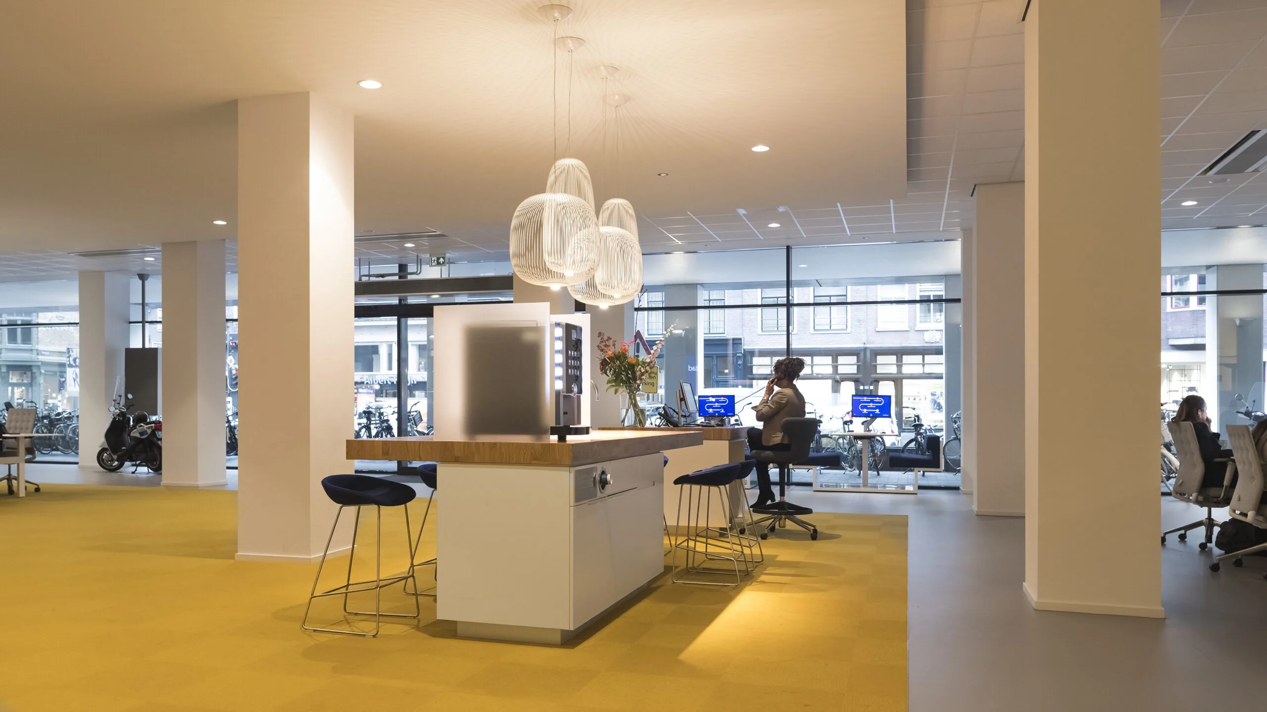 Moderne kantoorruimte aan de Vijzelstraat met gele vloerbedekking, open werkplekken en een centrale koffiecorner.