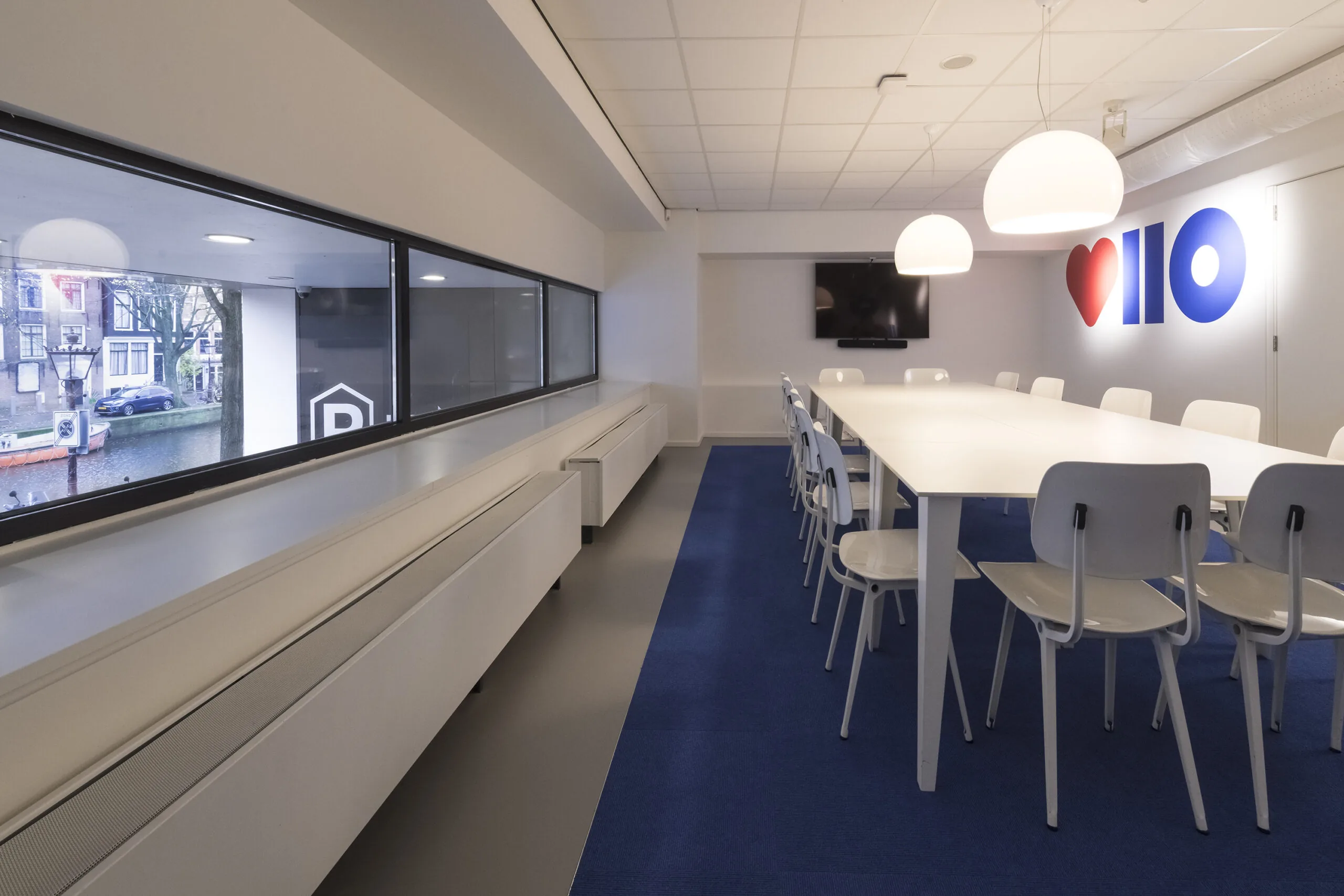 Vergaderruimte aan de Vijzelstraat met lange witte tafel, moderne stoelen, hanglampen en muurdecoratie met hartje en "110".