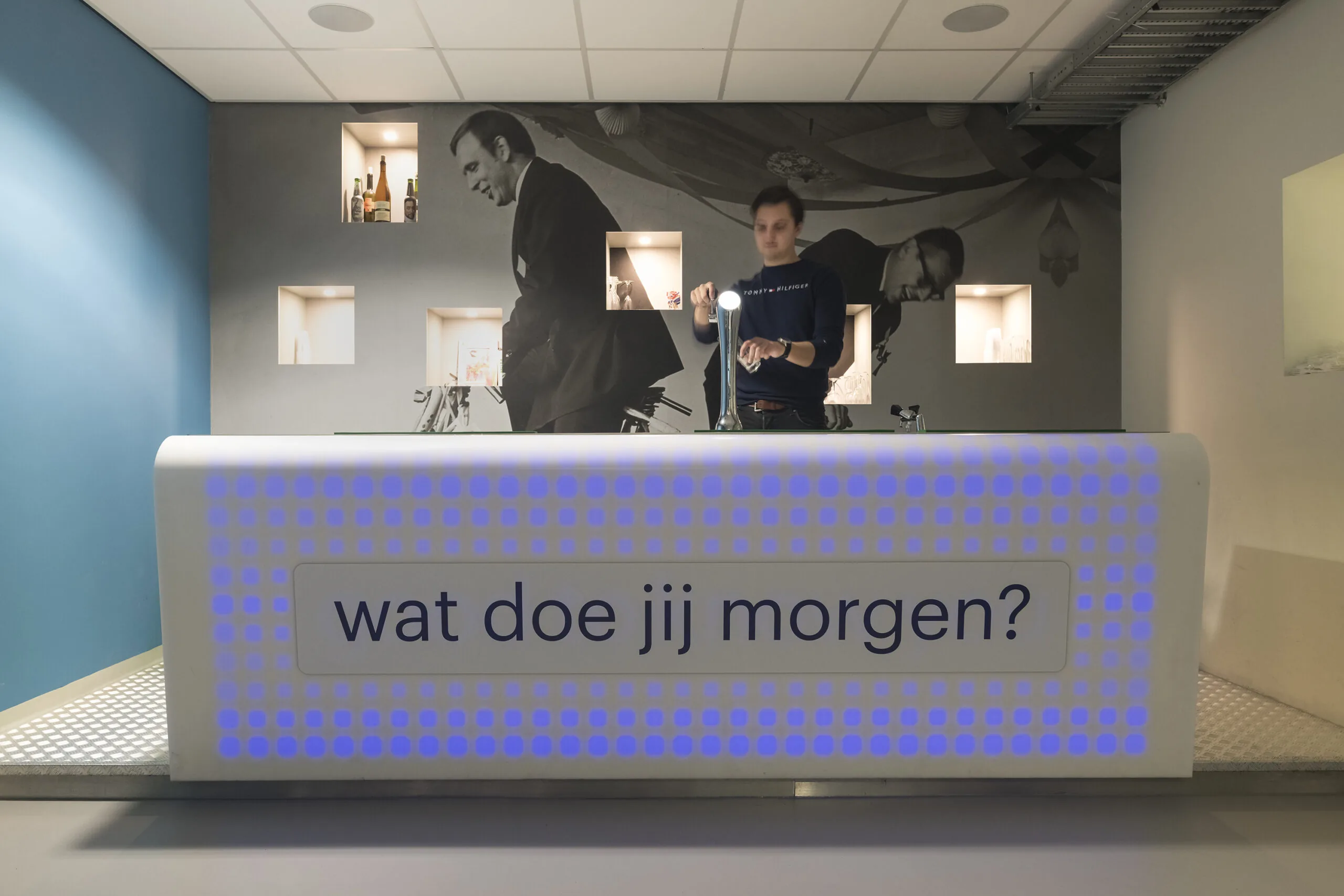 Man staat achter een verlichte balie met de tekst "wat doe jij morgen?" in een modern interieur met retro muurfoto's.