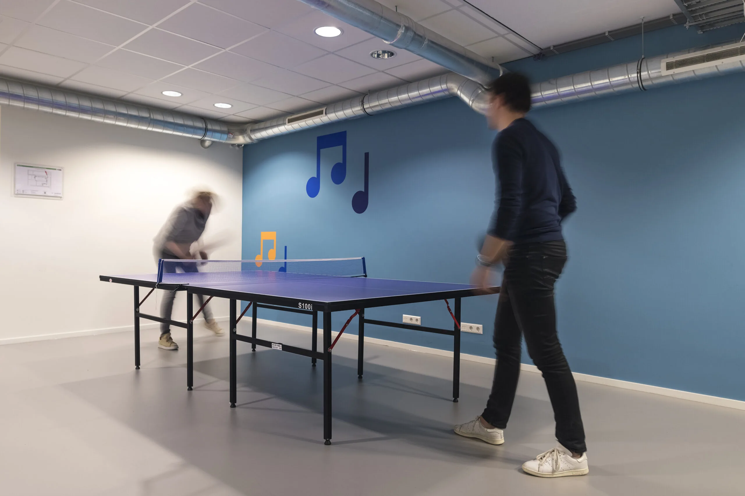 Twee personen spelen een potje tafeltennis in een modern kantoor met muzieknoten op de muur.