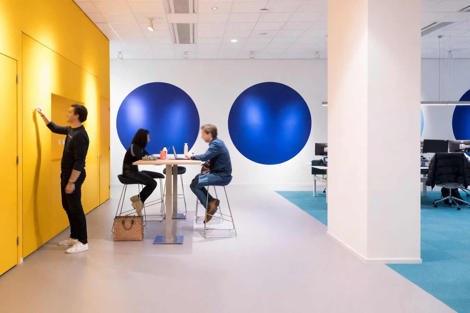 Moderne kantoorruimte op de Vijzelstraat met mensen die samenwerken aan een hoge tafel en een persoon die schrijft op een gele muur.