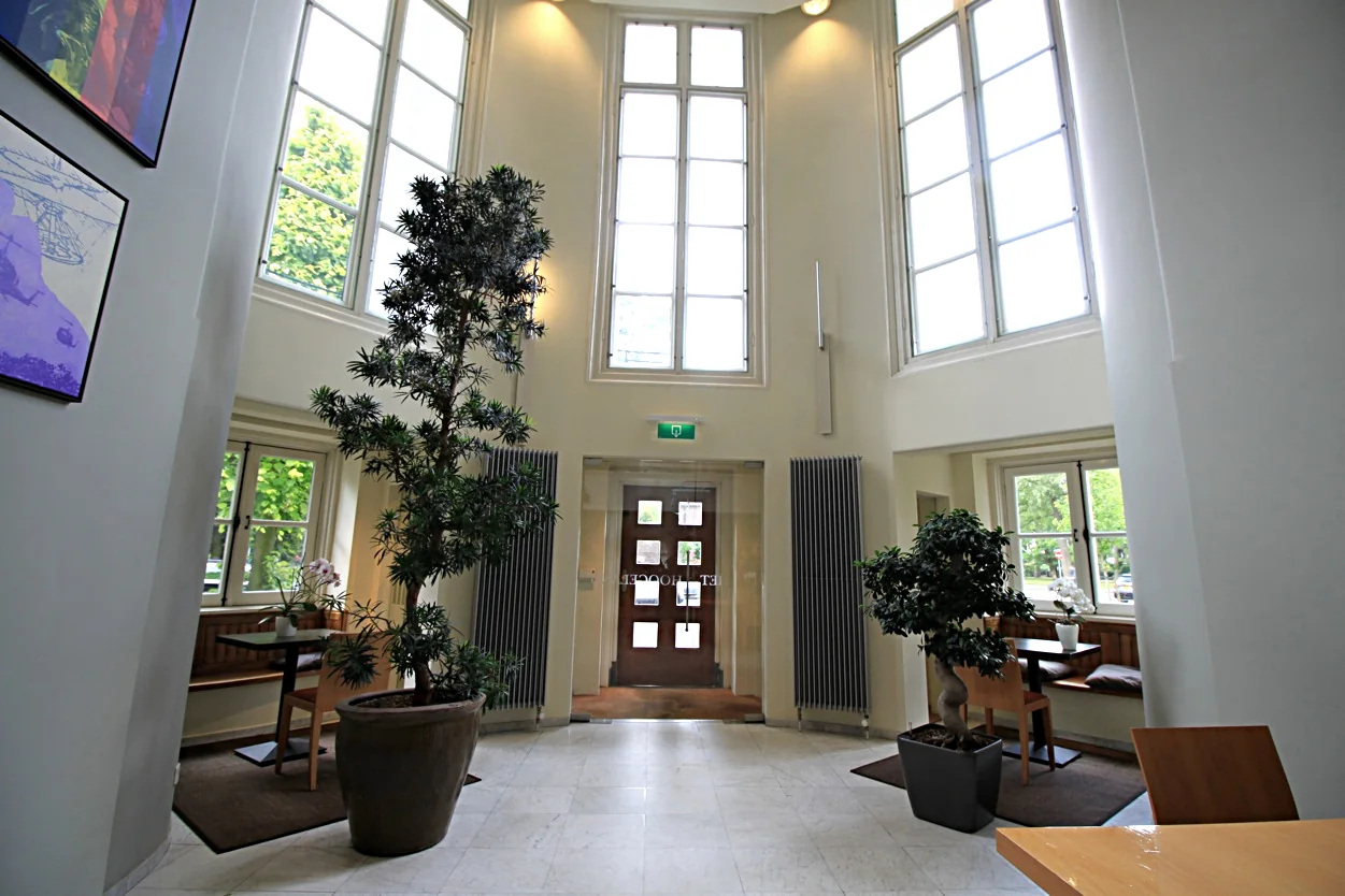 Lichte en hoge entreehal met grote ramen, planten en zithoeken in Museumlaan.
