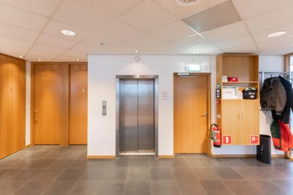 Hal van een kantoor of openbaar gebouw met een lift, meerdere deuren, kapstok en brandveiligheidsvoorzieningen.