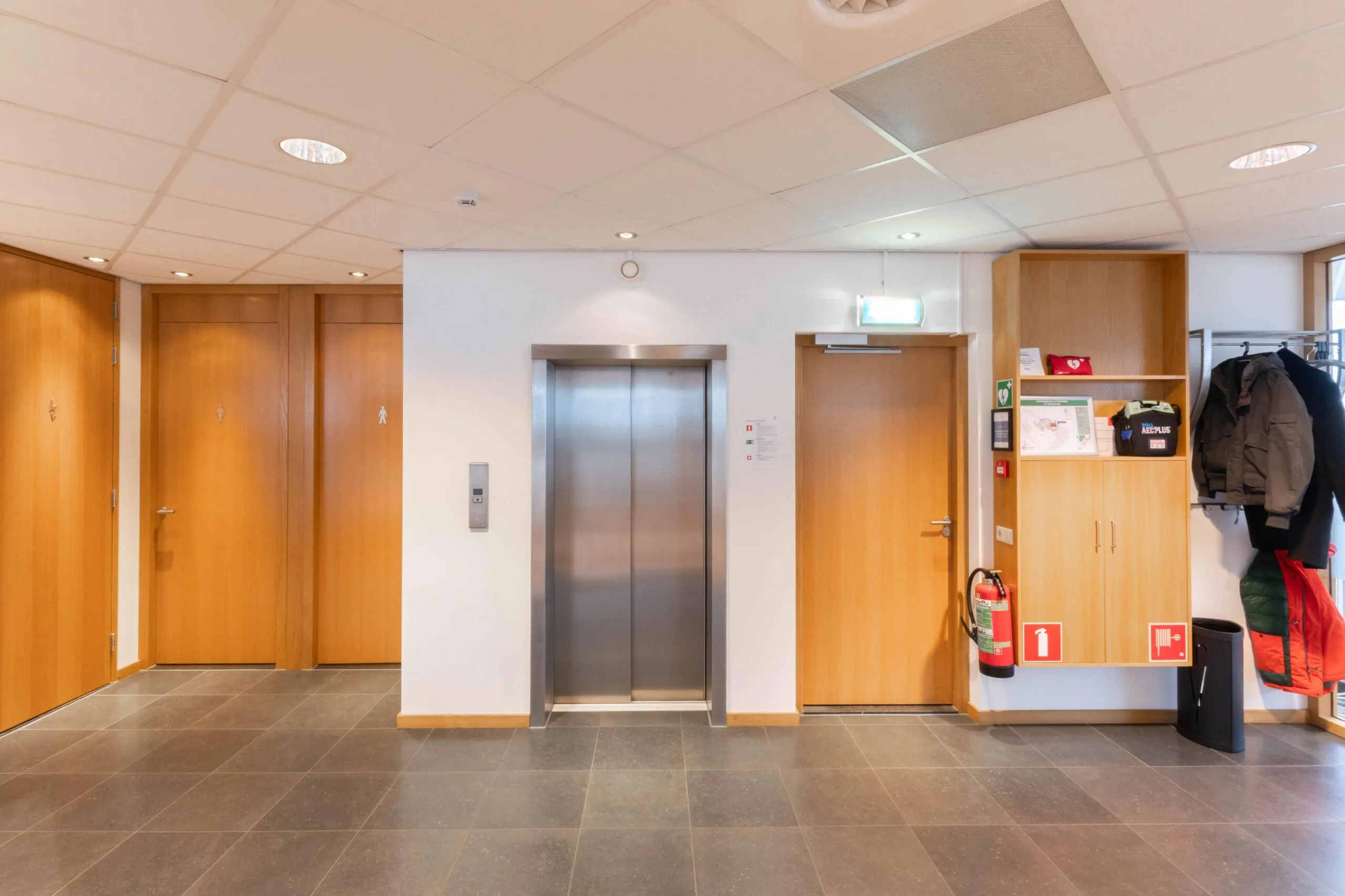 Hal van een kantoor of openbaar gebouw met een lift, meerdere deuren, kapstok en brandveiligheidsvoorzieningen.