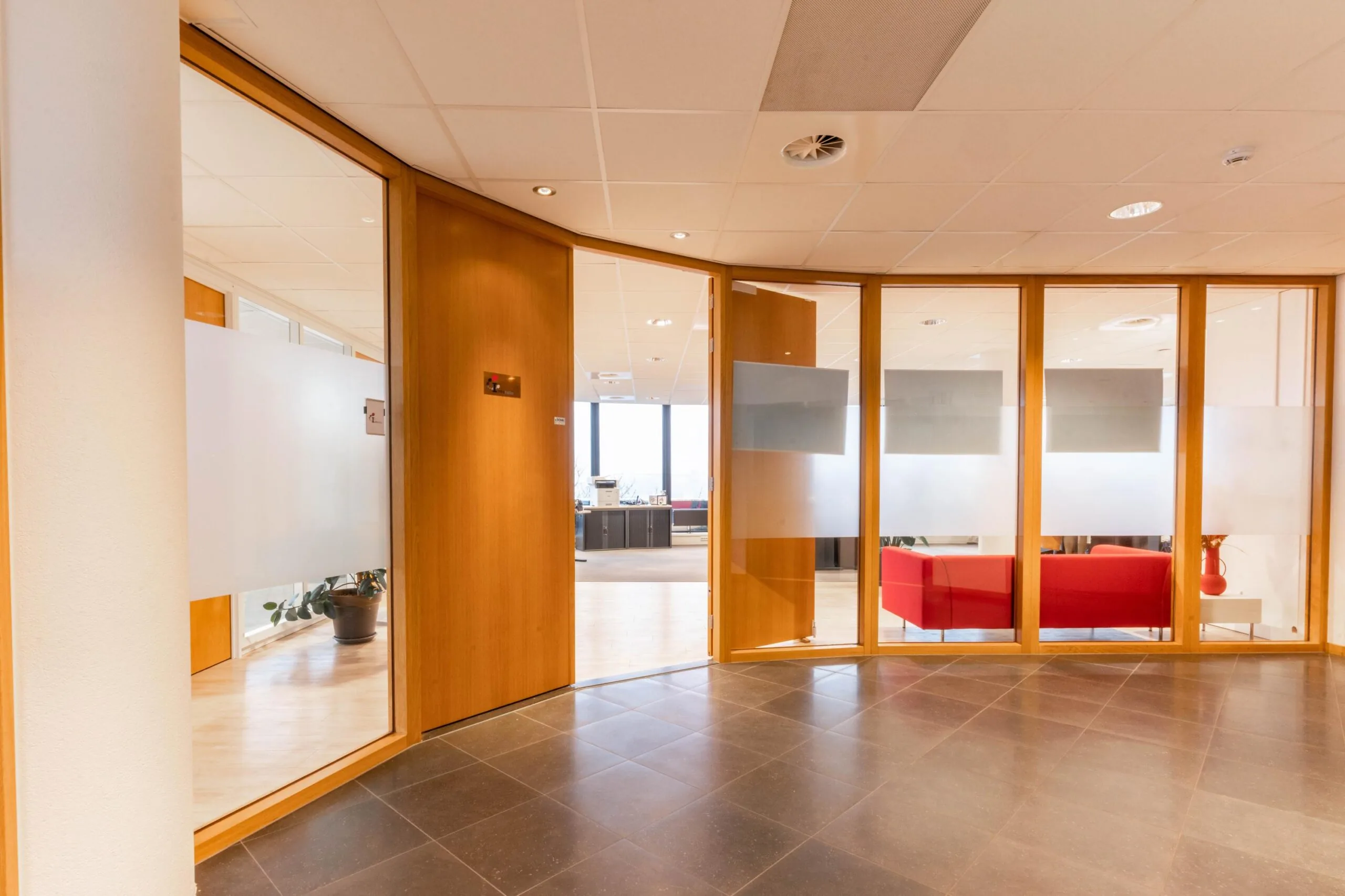 Ingang van een modern kantoor aan de 1e Hogeweg met glazen wanden, houten kozijnen en een rode zithoek.