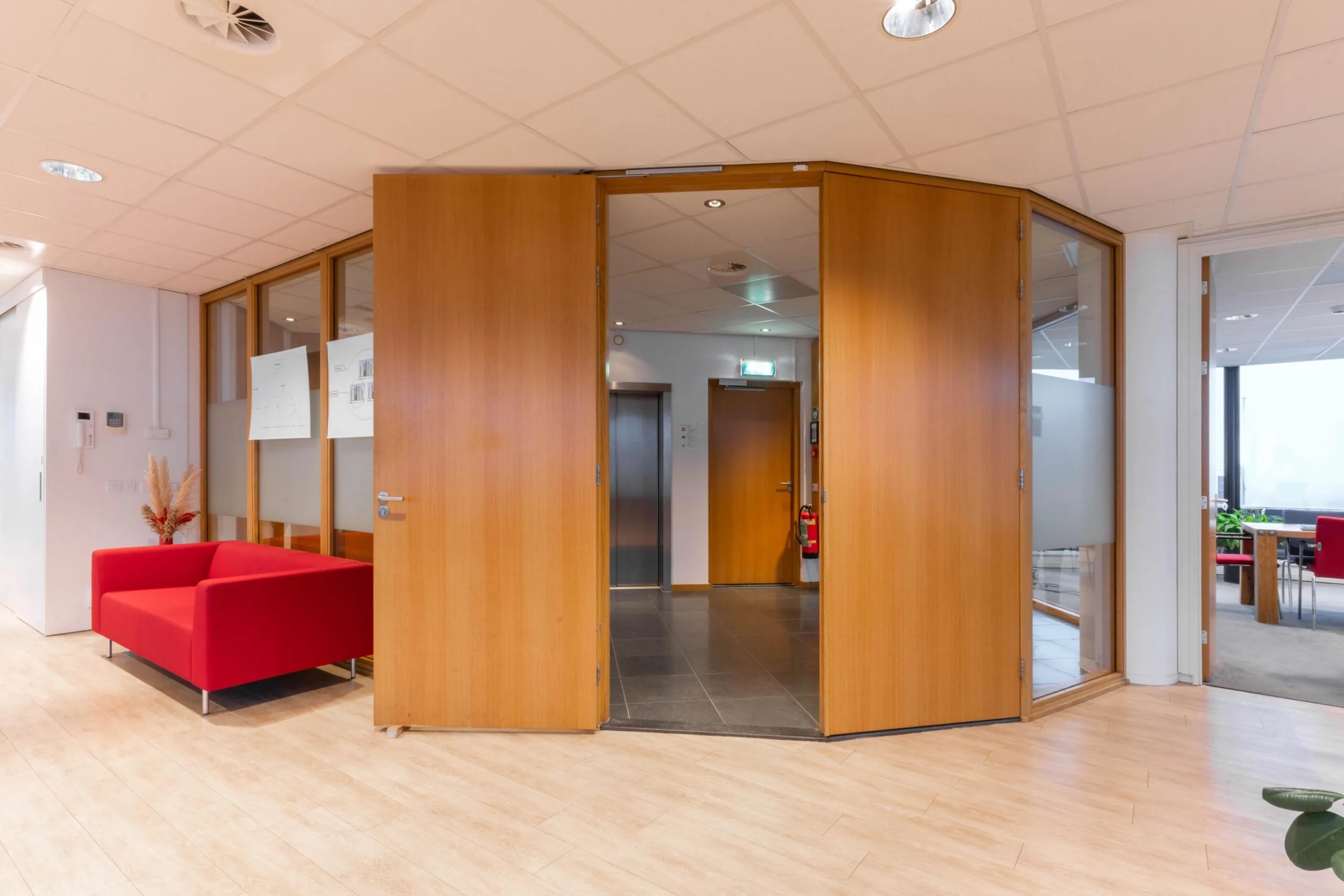 Ingang van een modern kantoorpand aan de 1e Hogeweg met houten deuren, glazen wanden en een rode wachtbank.