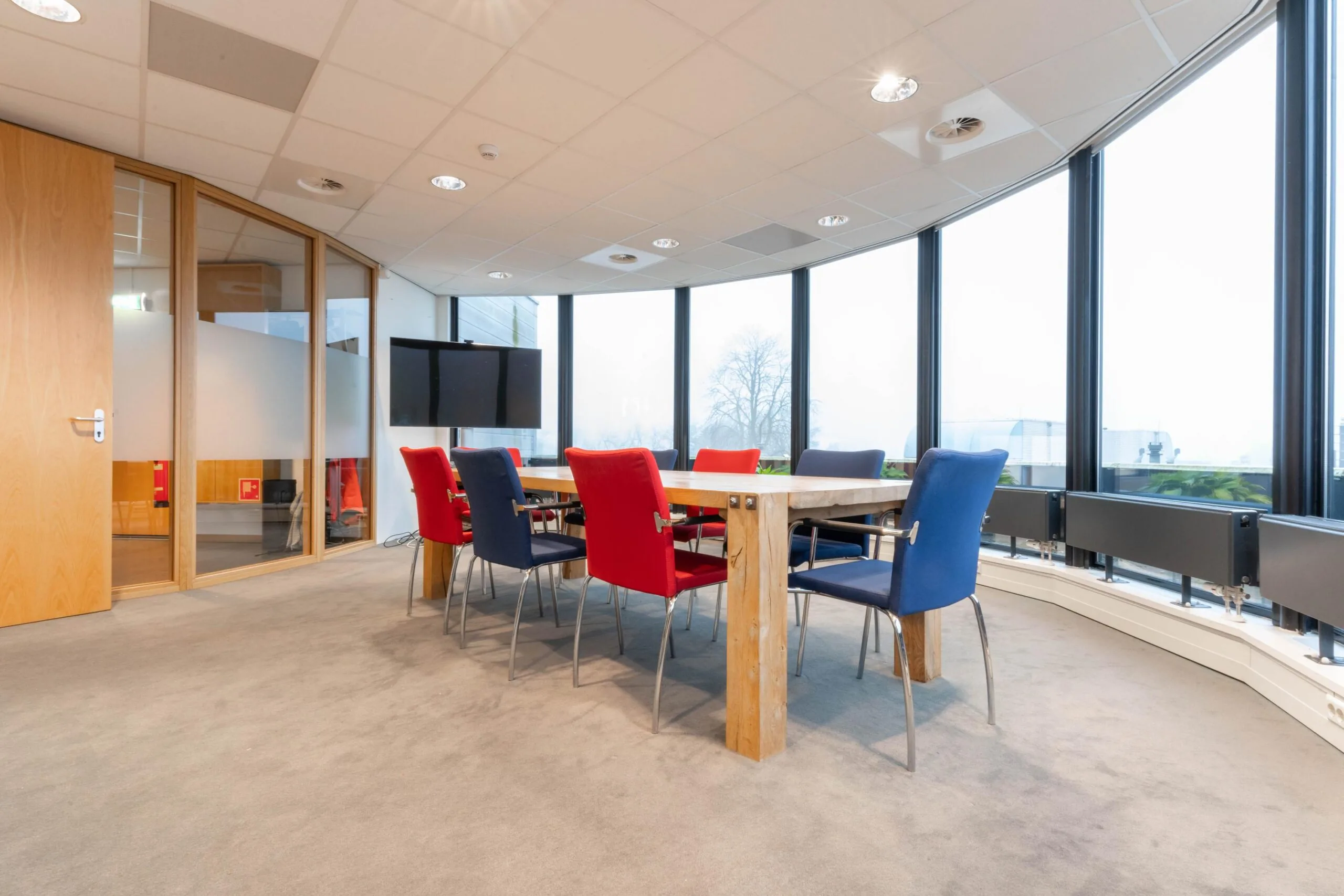Vergaderruimte met houten tafel, kleurrijke stoelen en grote ramen aan de 1e Hogeweg.