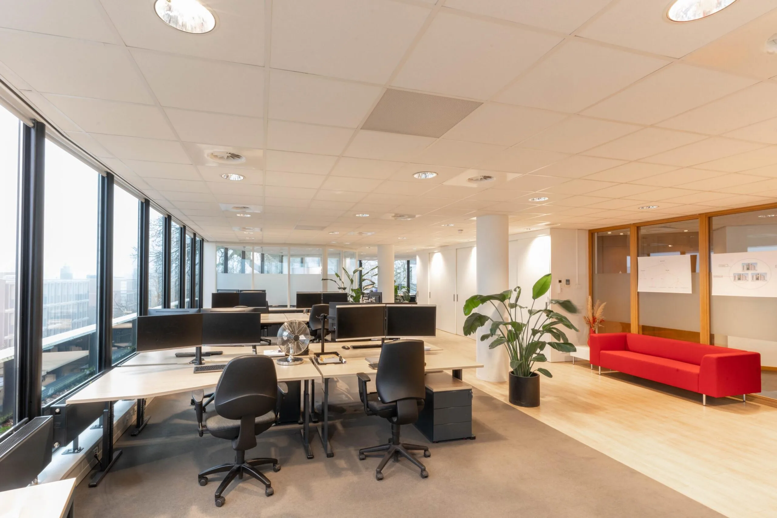 Moderne, open kantoorruimte aan de 1e Hogeweg met werkplekken, grote ramen en een rode bank in de ontvangstruimte.