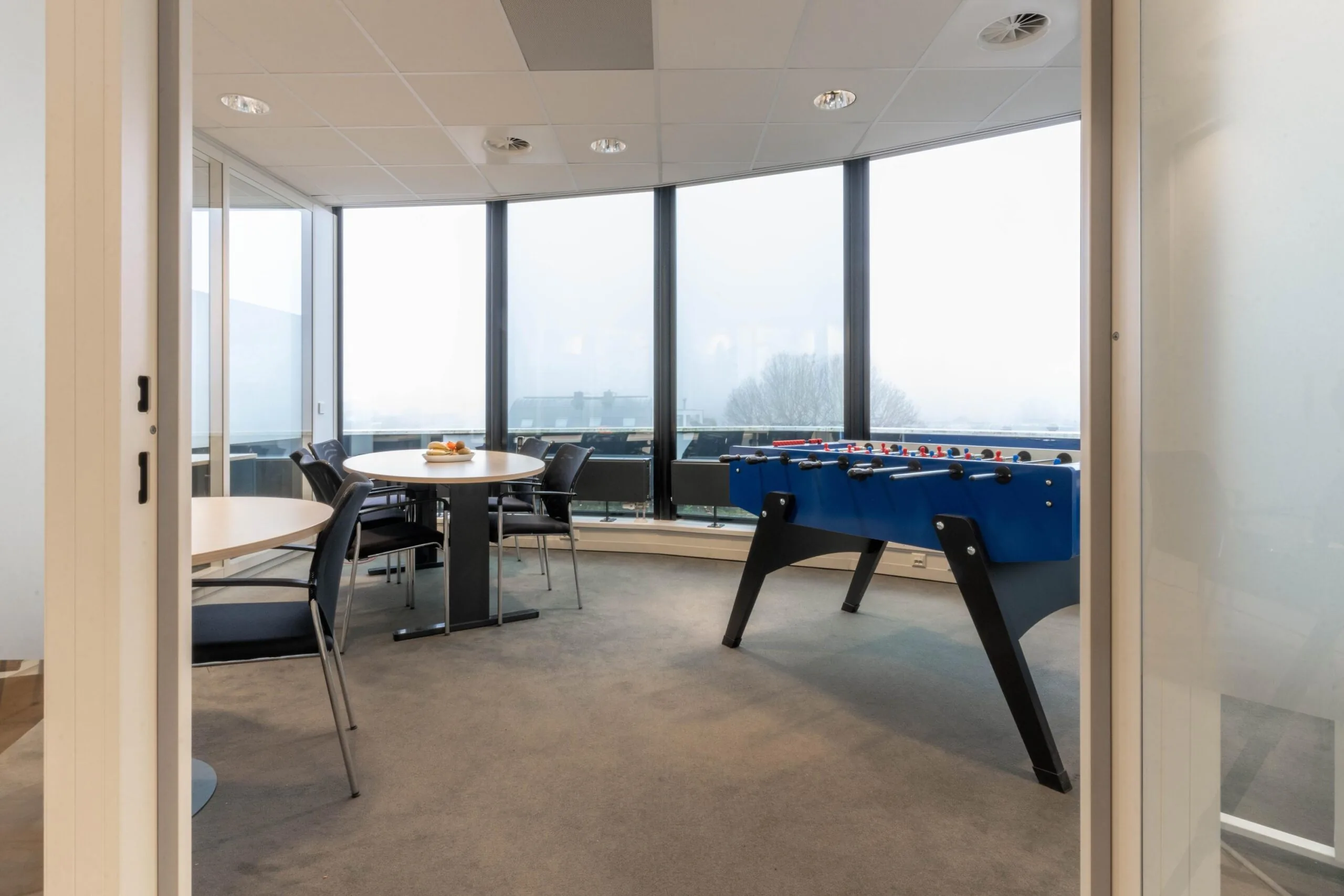 Kantineruimte met ronde tafels, stoelen en een blauwe tafelvoetbaltafel bij grote ramen in een modern kantoorpand aan de 1e Hogeweg.