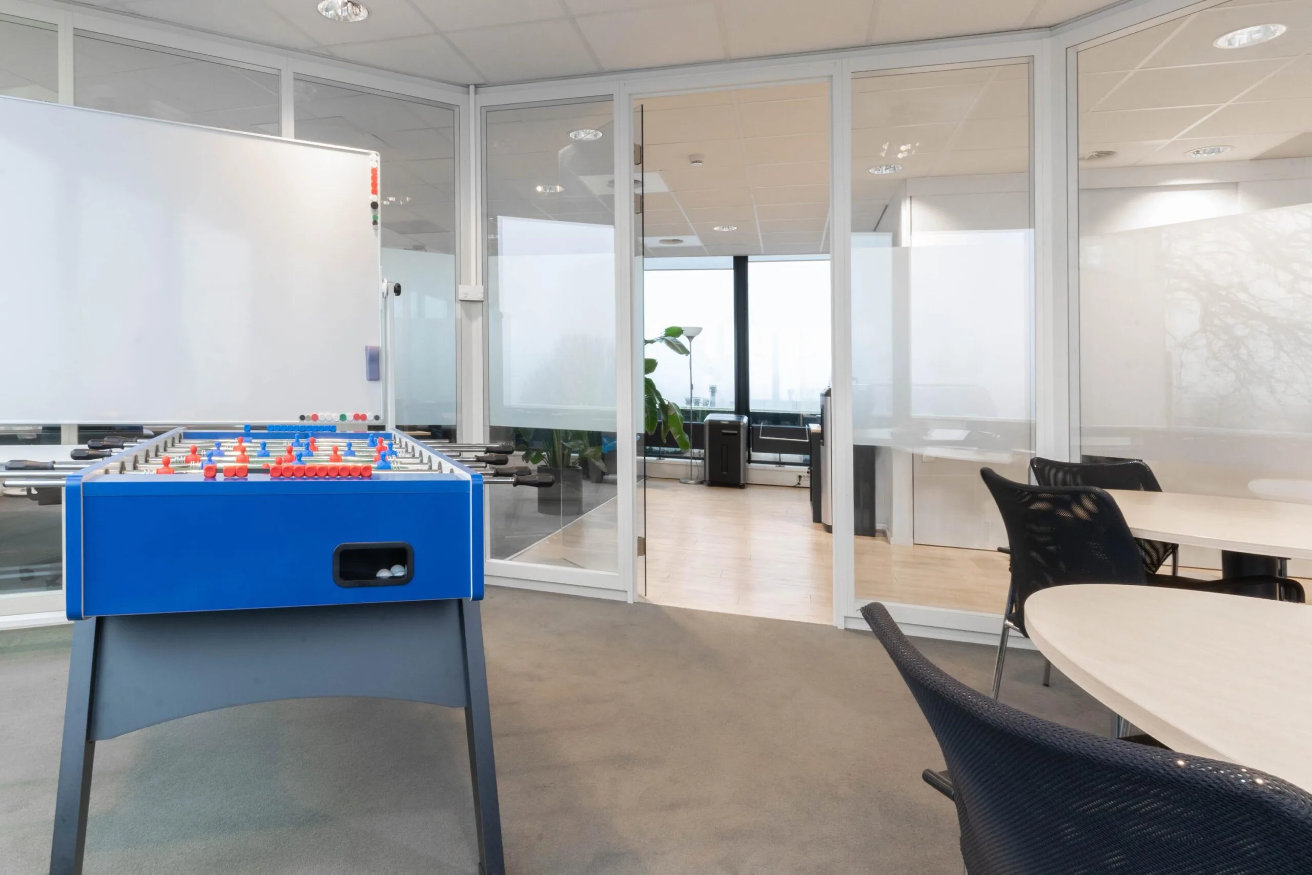 Moderne kantoorruimte met glazen wanden, een tafelvoetbaltafel, whiteboard en ronde vergadertafels.