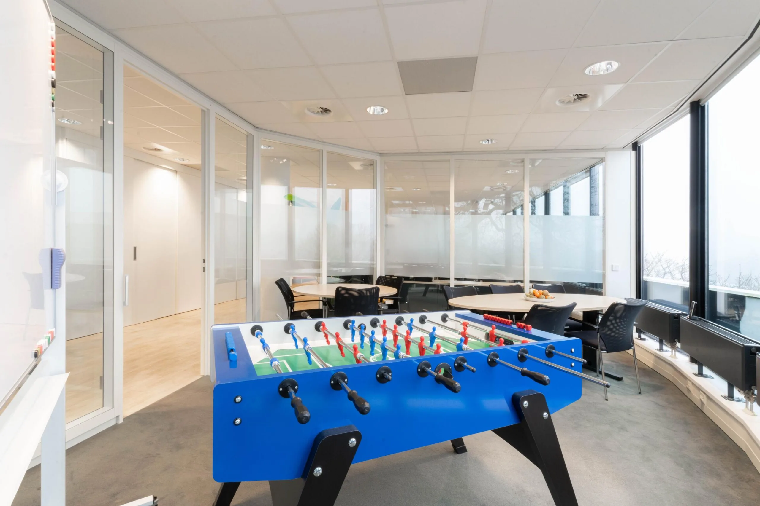 Moderne kantoorruimte met grote ramen, een blauwe tafelvoetbaltafel en meerdere zithoeken.