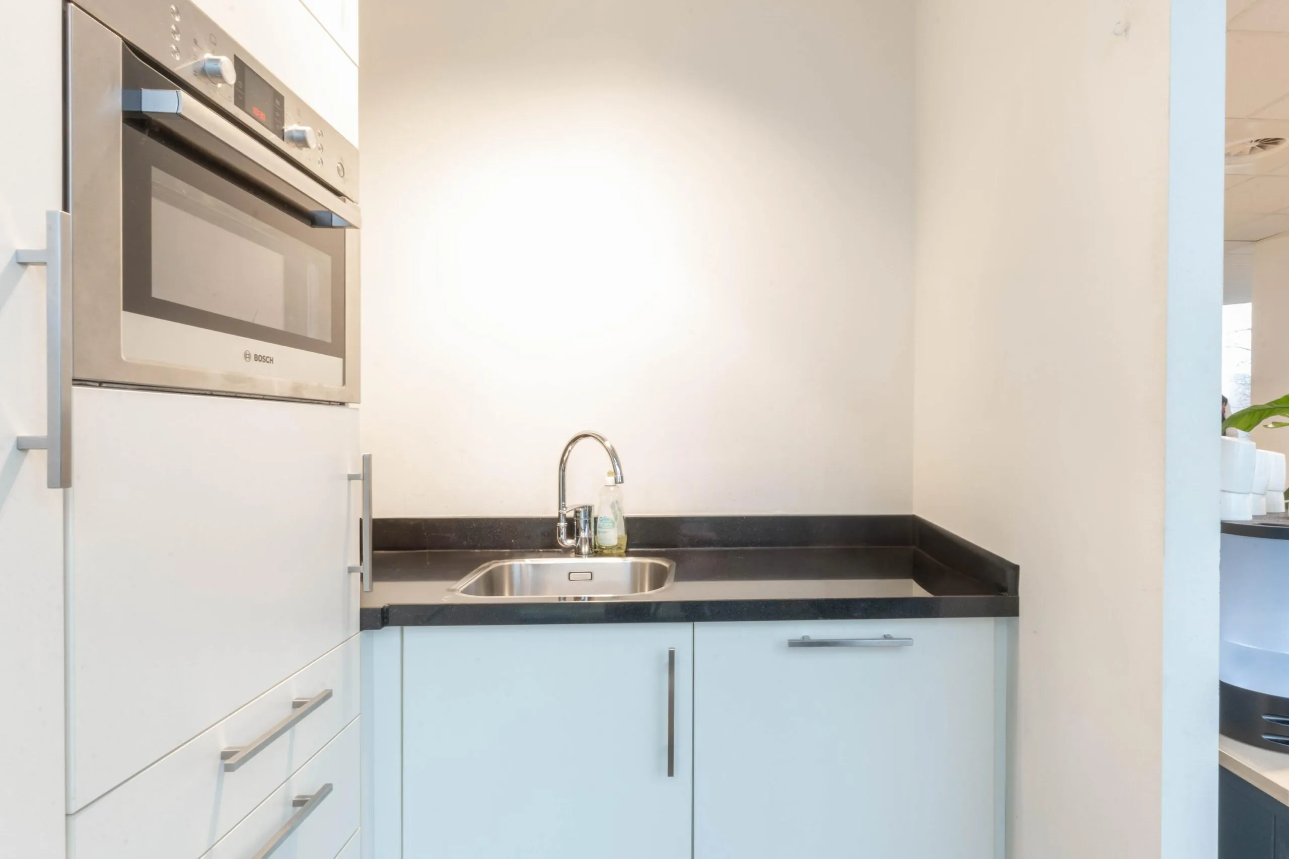 Kleine, moderne kitchenette met een ingebouwde Bosch oven, rvs-wastafel en witte kasten.