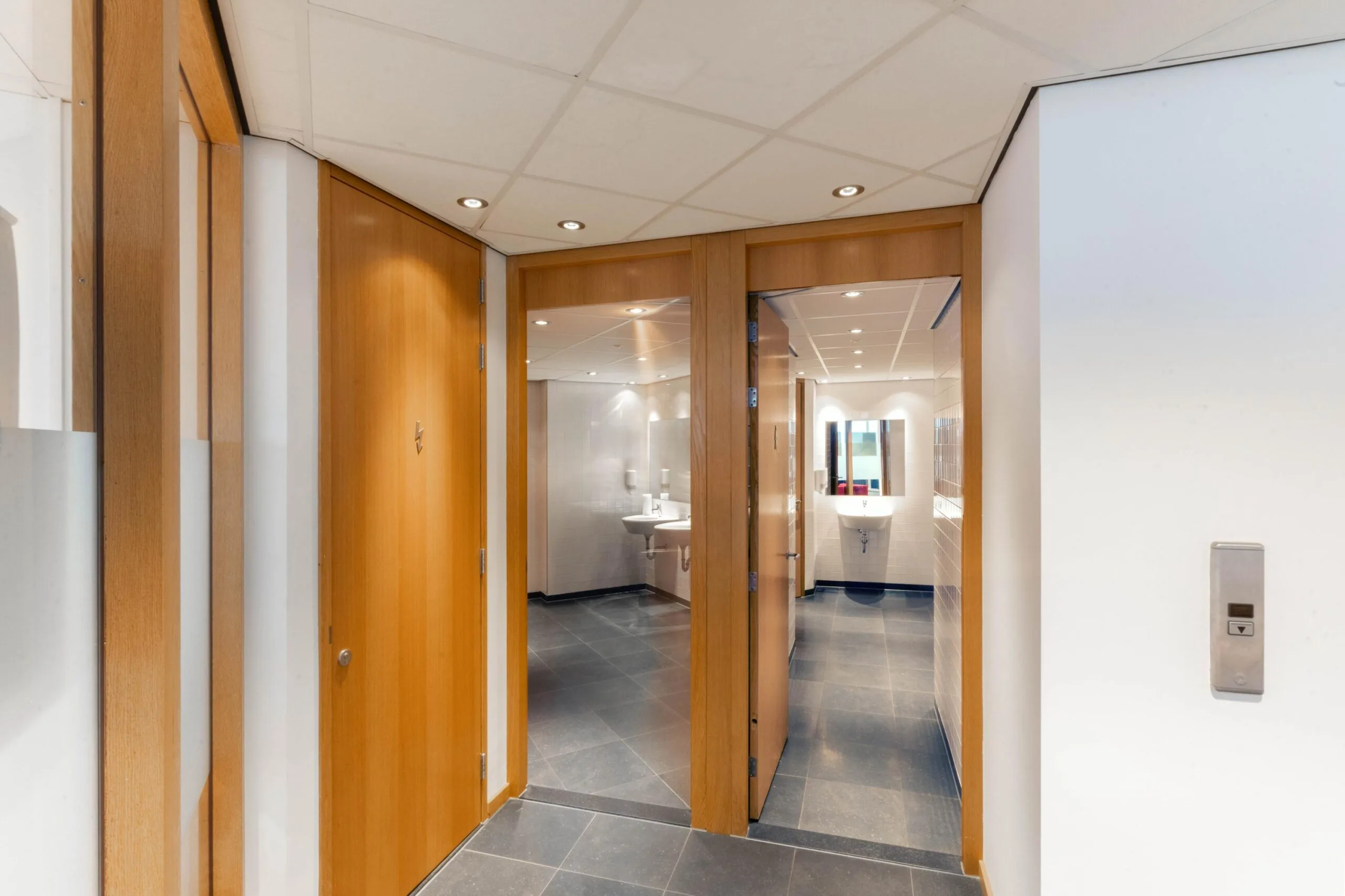 Ingang van een modern sanitairgedeelte met houten deuren en wastafels in een nette, betegelde ruimte.