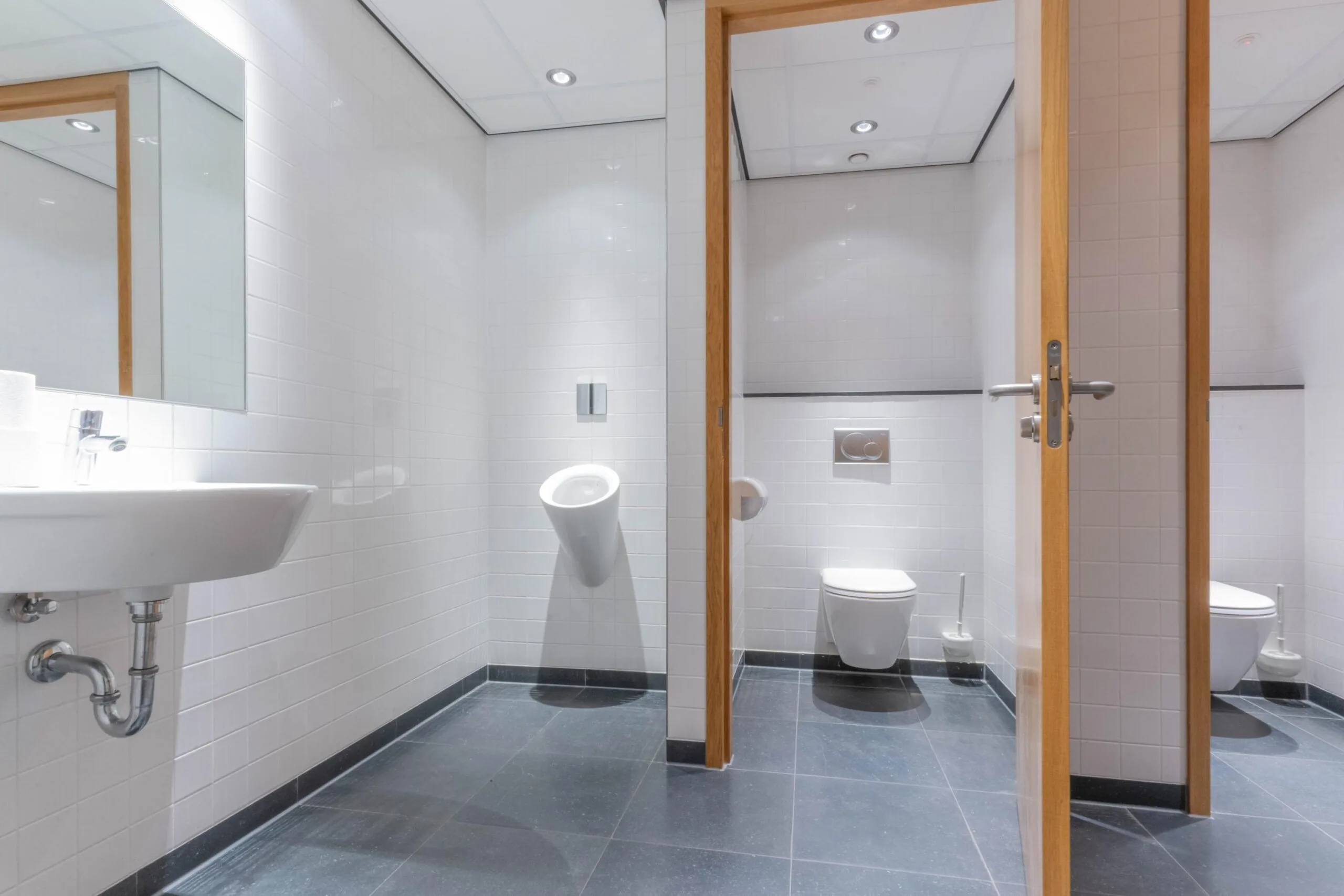 Moderne, wit betegelde sanitaire ruimte met urinoir, twee toiletten en een wastafel in 1e Hogeweg.