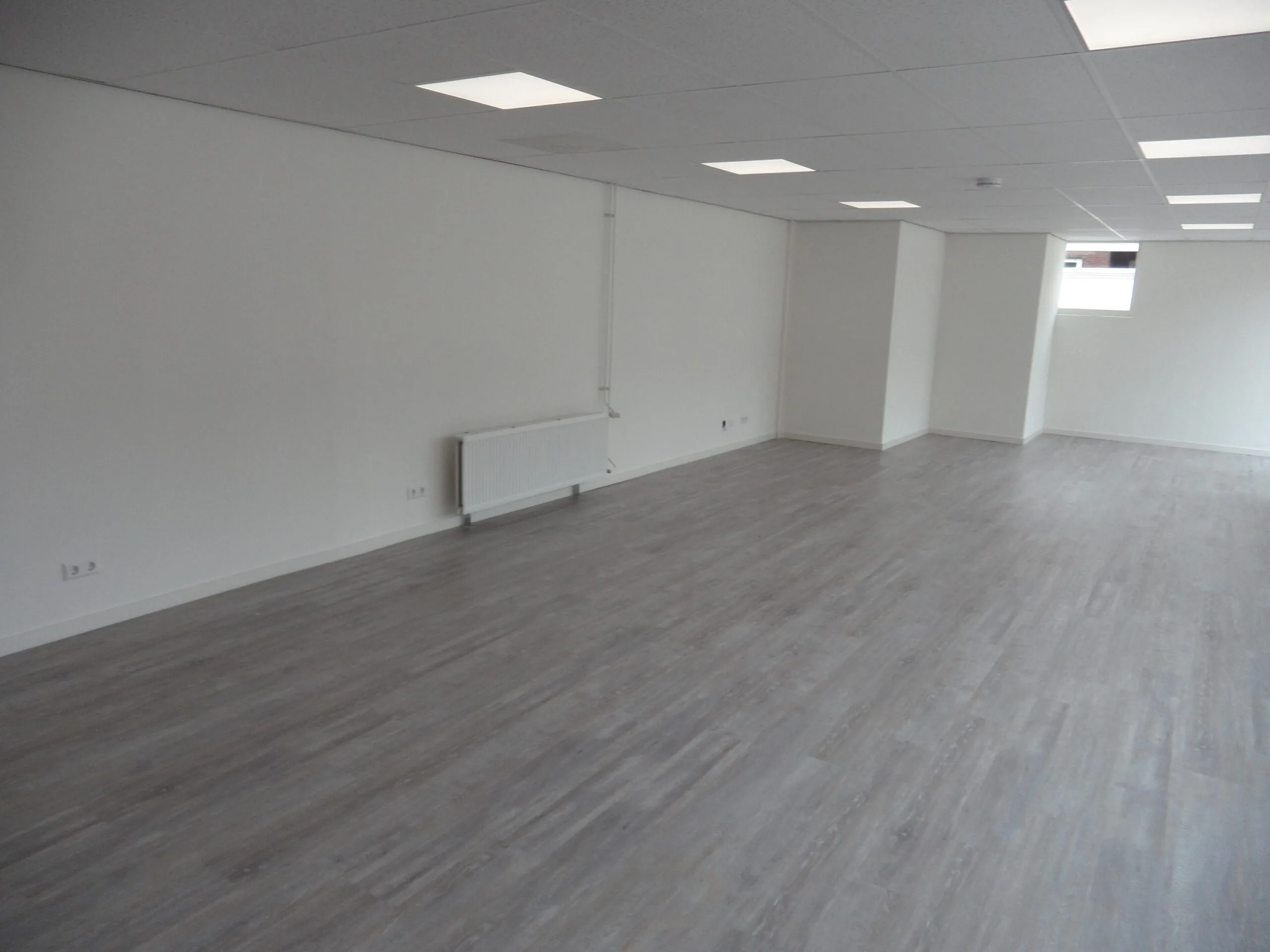 Lege, strak afgewerkte ruimte met grijze vloer, witte muren en systeemplafond aan de Jacques Perkstraat.