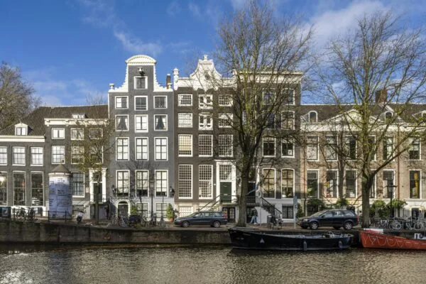 Gevels van historische grachtenpanden aan de Herengracht in Amsterdam met bomen, geparkeerde auto's en boten op de voorgrond.