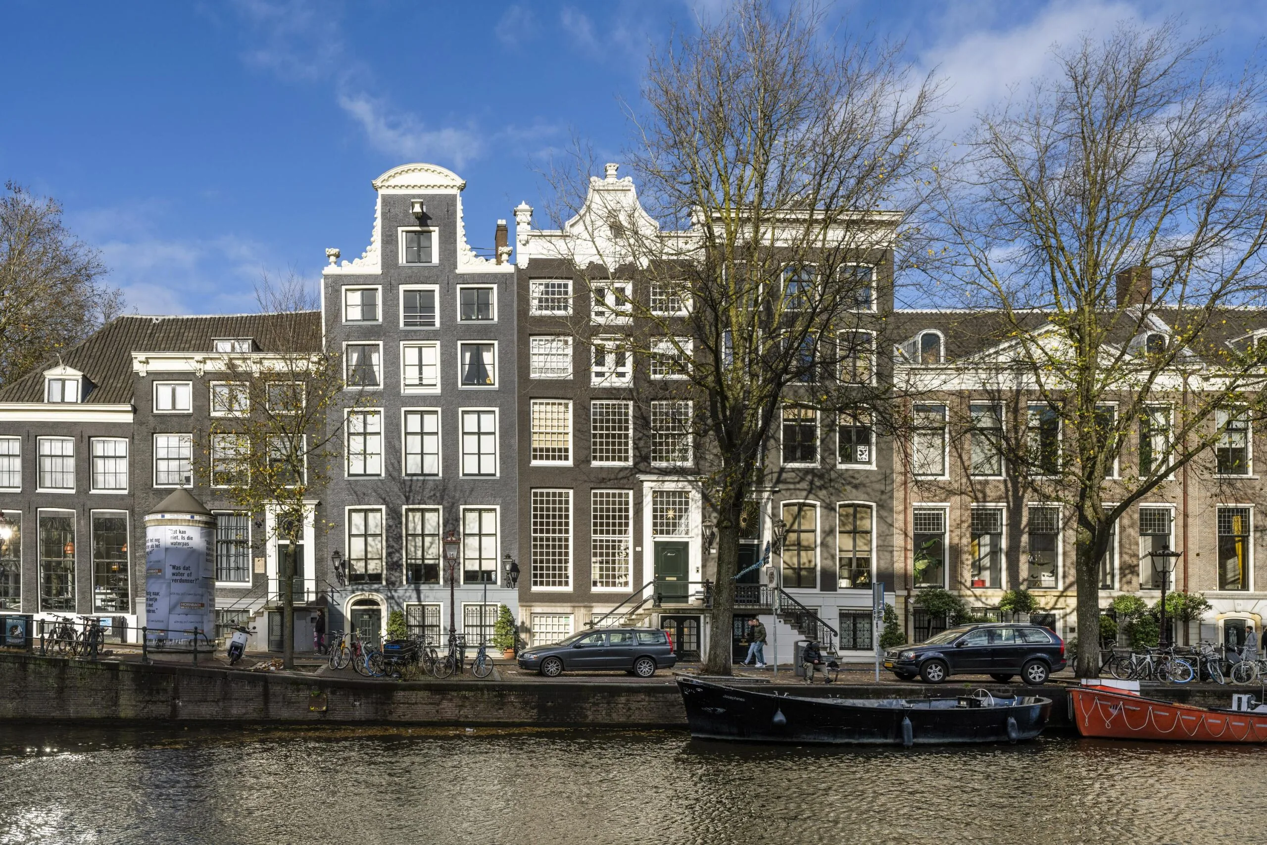 Gevels van historische grachtenpanden aan de Herengracht in Amsterdam met bomen, geparkeerde auto's en boten op de voorgrond.