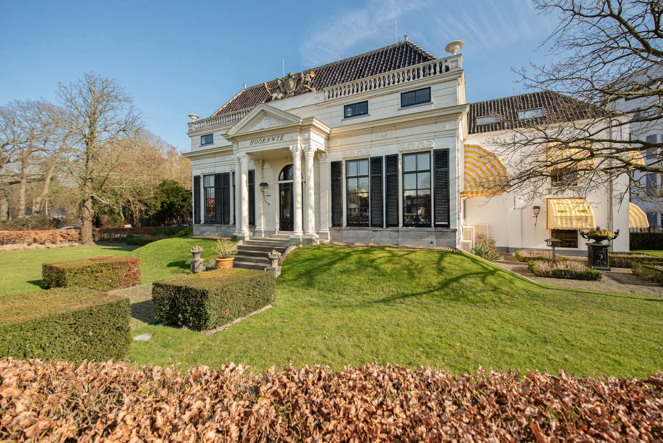 Witte klassieke villa met zuilen en de naam 'Hoornwyck' aan de Jan Thijssenweg, omgeven door een verzorgde tuin.