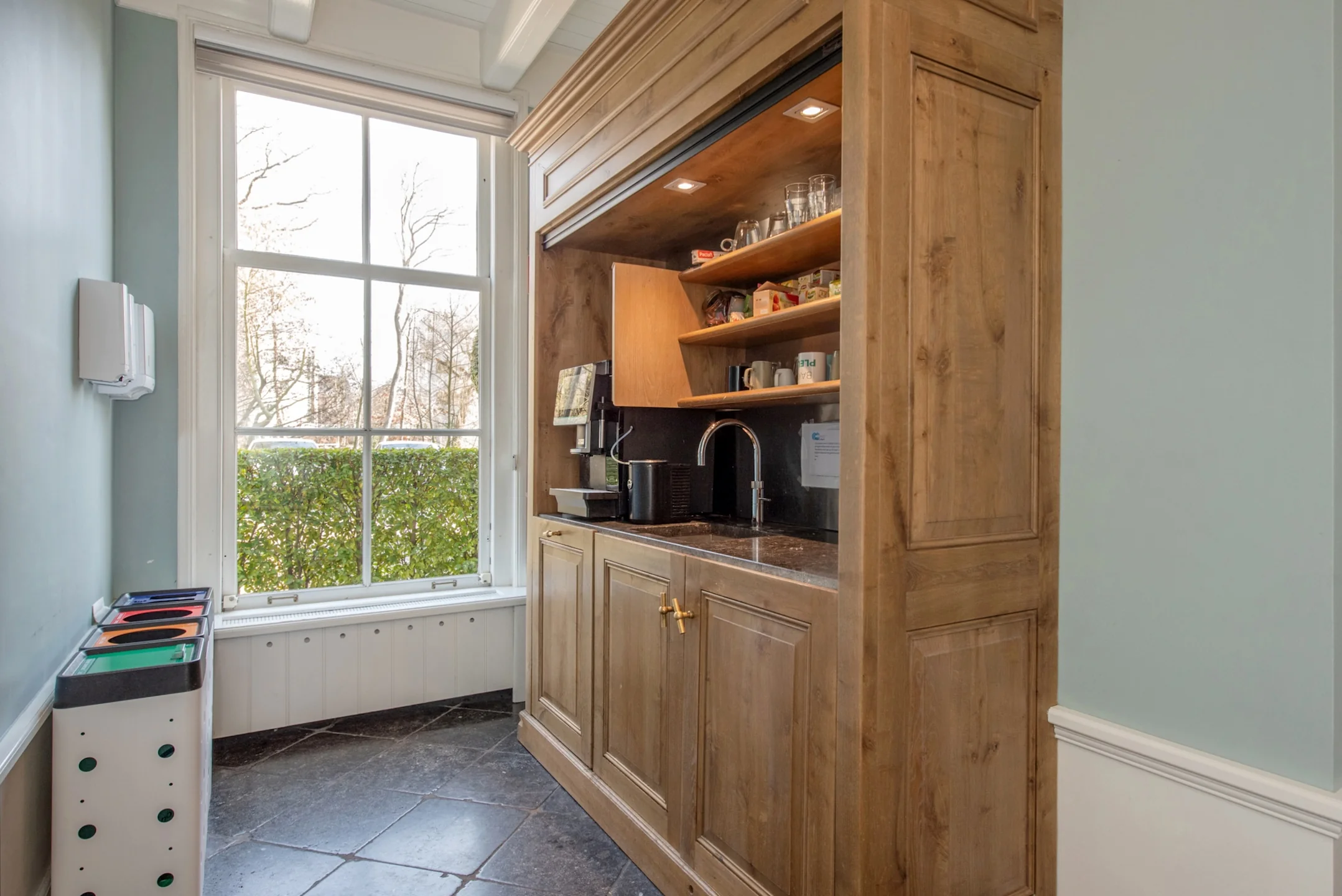 Compacte kitchenette met houten kast, ingebouwde wasbak, koffiezetapparaat en opbergrekken, gelegen naast een groot raam.