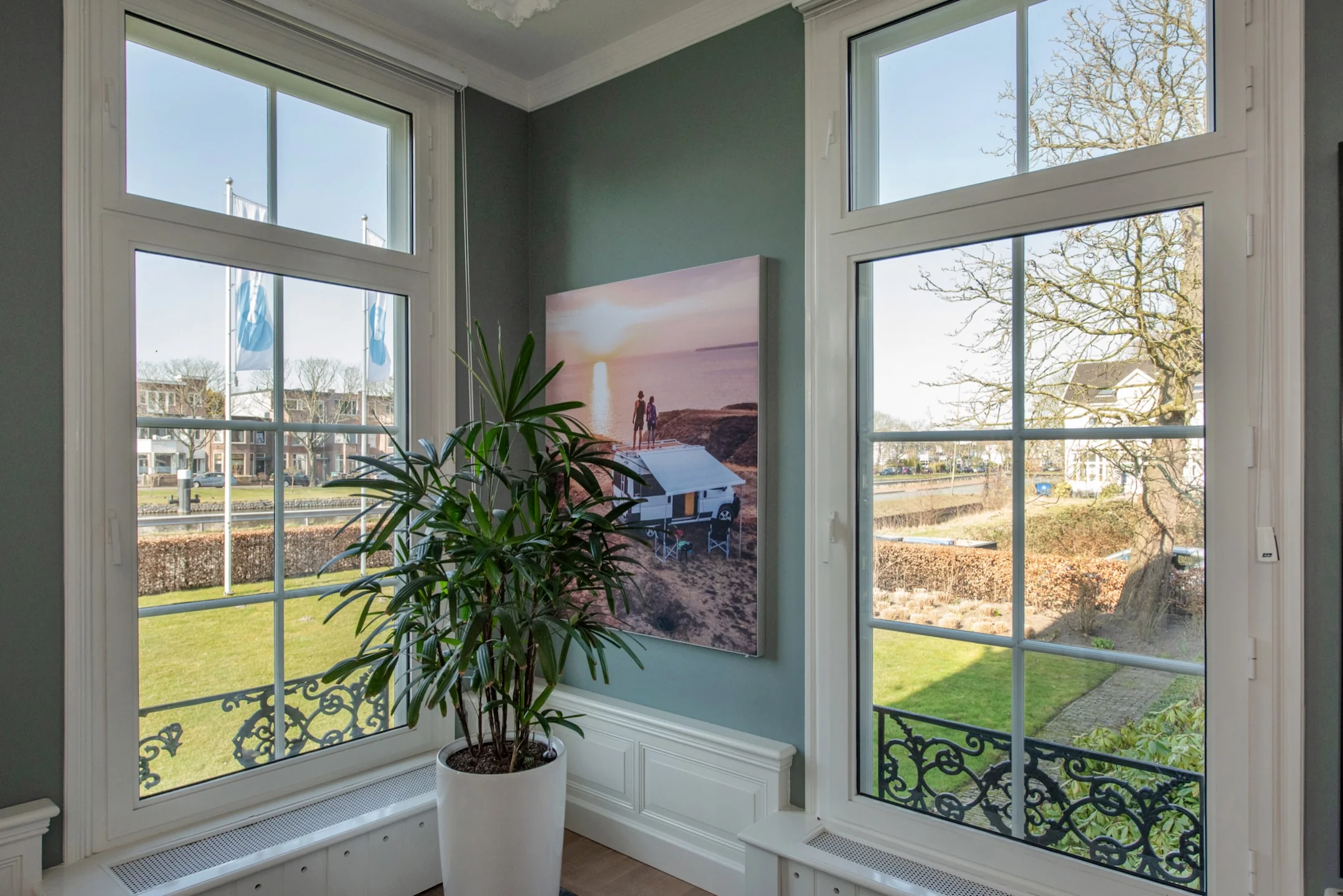 Binnenhoek van een kamer met grote ramen, een groene plant in een witte pot en een wandfoto van een stel bij een caravan in de avondzon.