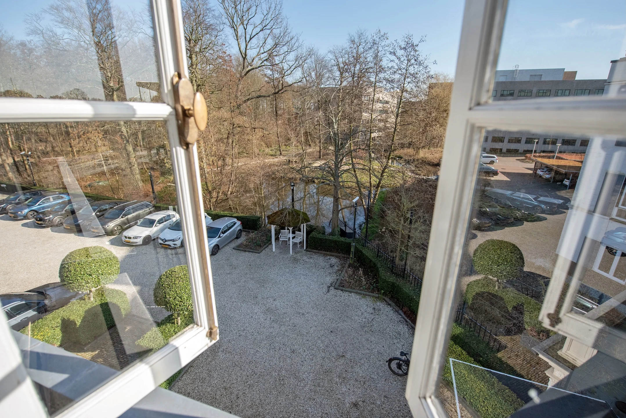 Uitzicht vanuit een open raam op de Jan Thijssenweg, met geparkeerde auto's, een tuinpaviljoen en bomen rond een vijver.