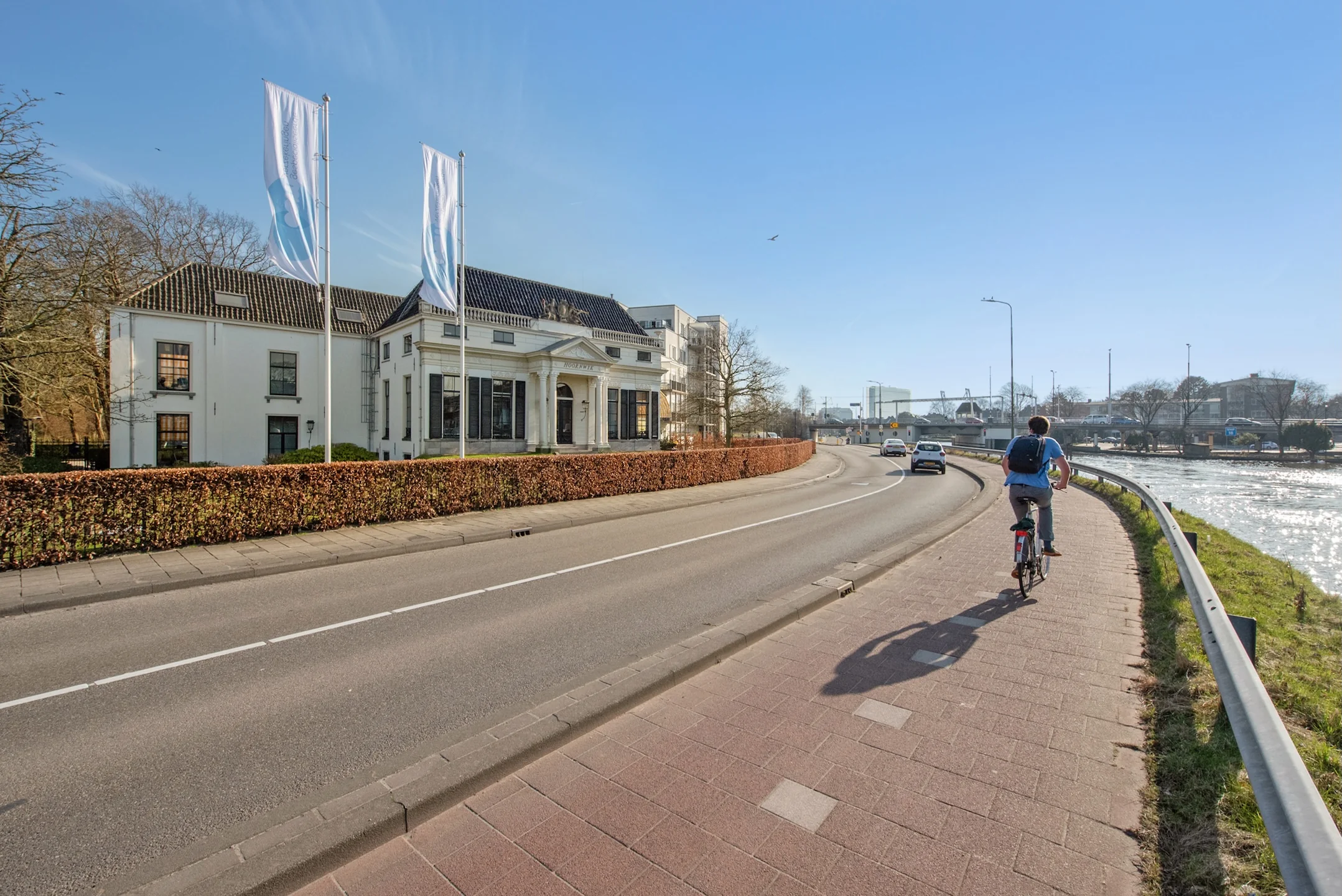 Fietser op het fietspad langs de Jan Thijssenweg met een klassiek wit gebouw en de rivier aan de rechterzijde.