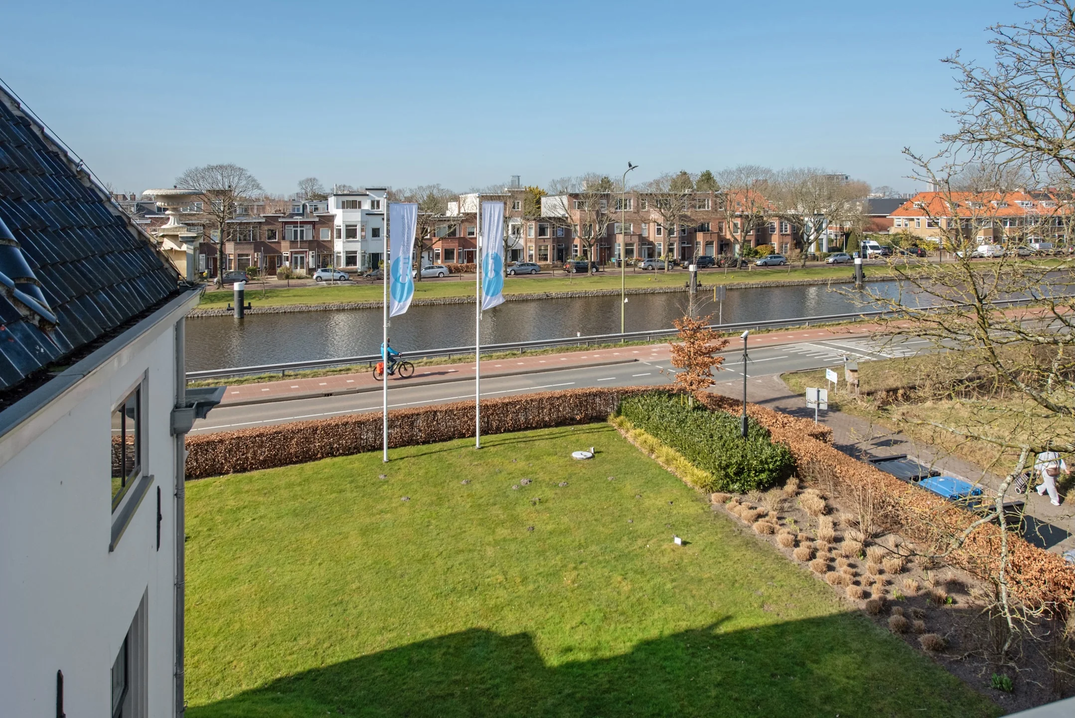 Uitzicht vanaf een gebouw aan de Jan Thijssenweg op een gracht, woonhuizen en een groen gazon met vlaggen.