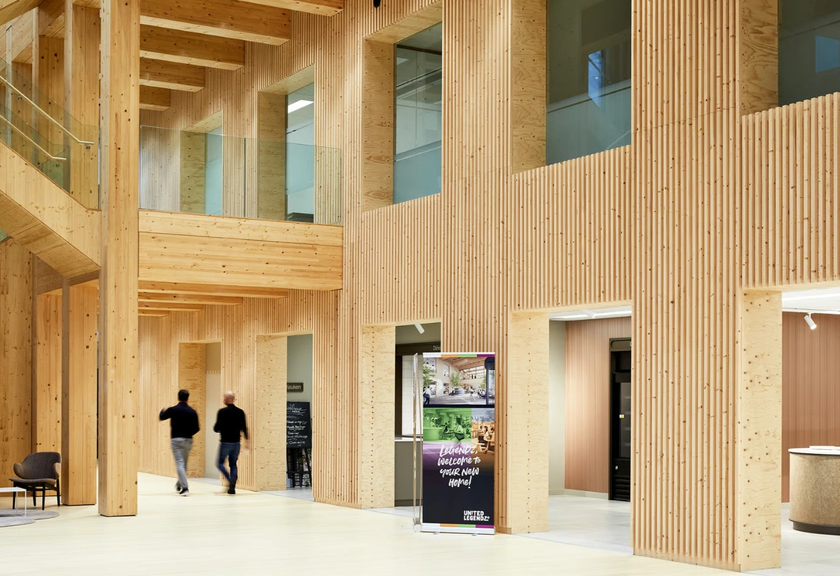 Moderne houten entreehal van het gebouw aan de Basisweg met twee personen die door de gang lopen en een welkomstbord van United Legendz.