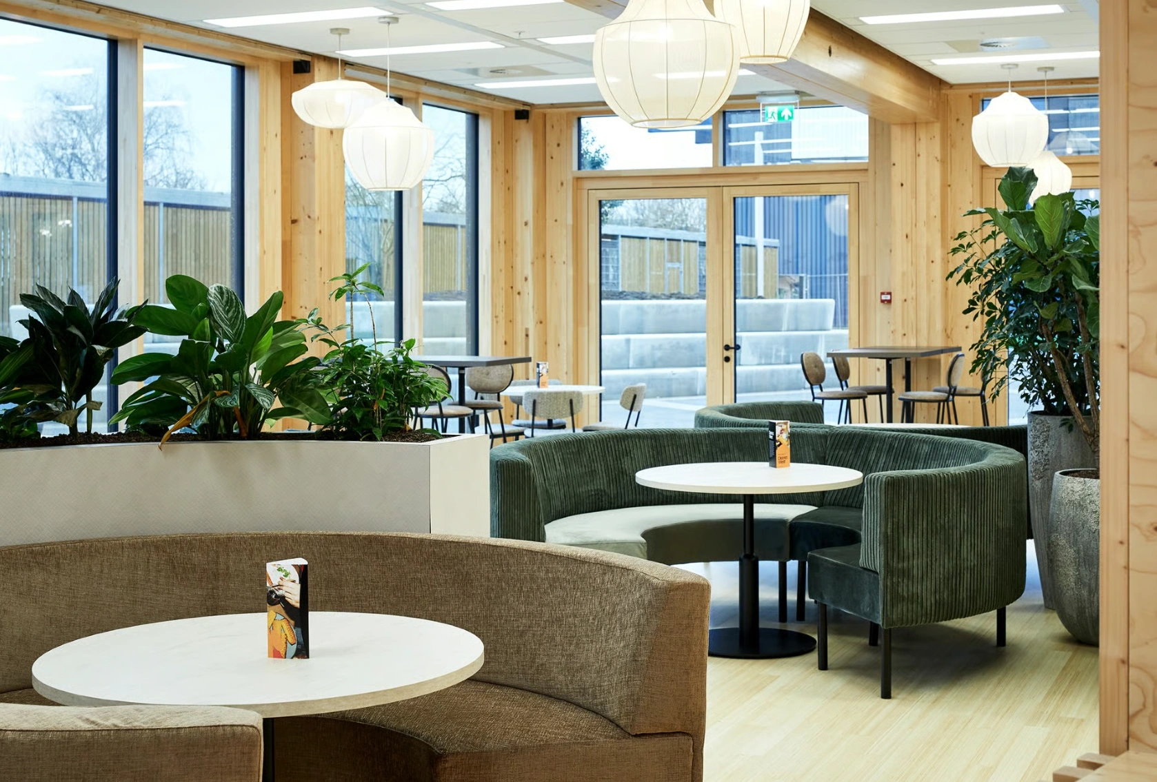 Moderne, lichte zithoek in een horecaruimte aan de Basisweg met ronde banken, planten en grote ramen.