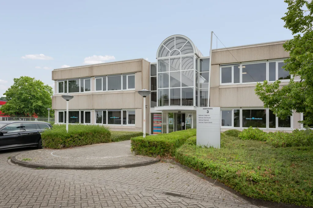 Vooraanzicht van een kantoorpand aan de Energieweg 3 met het logo van Veltman Uitgevers op de gevel en informatiebord in de tuin.