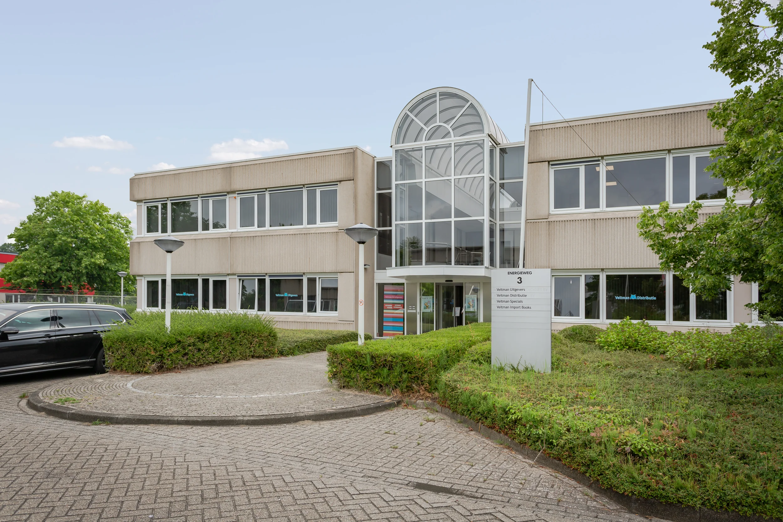 Vooraanzicht van een kantoorpand aan de Energieweg 3 met het logo van Veltman Uitgevers op de gevel en informatiebord in de tuin.