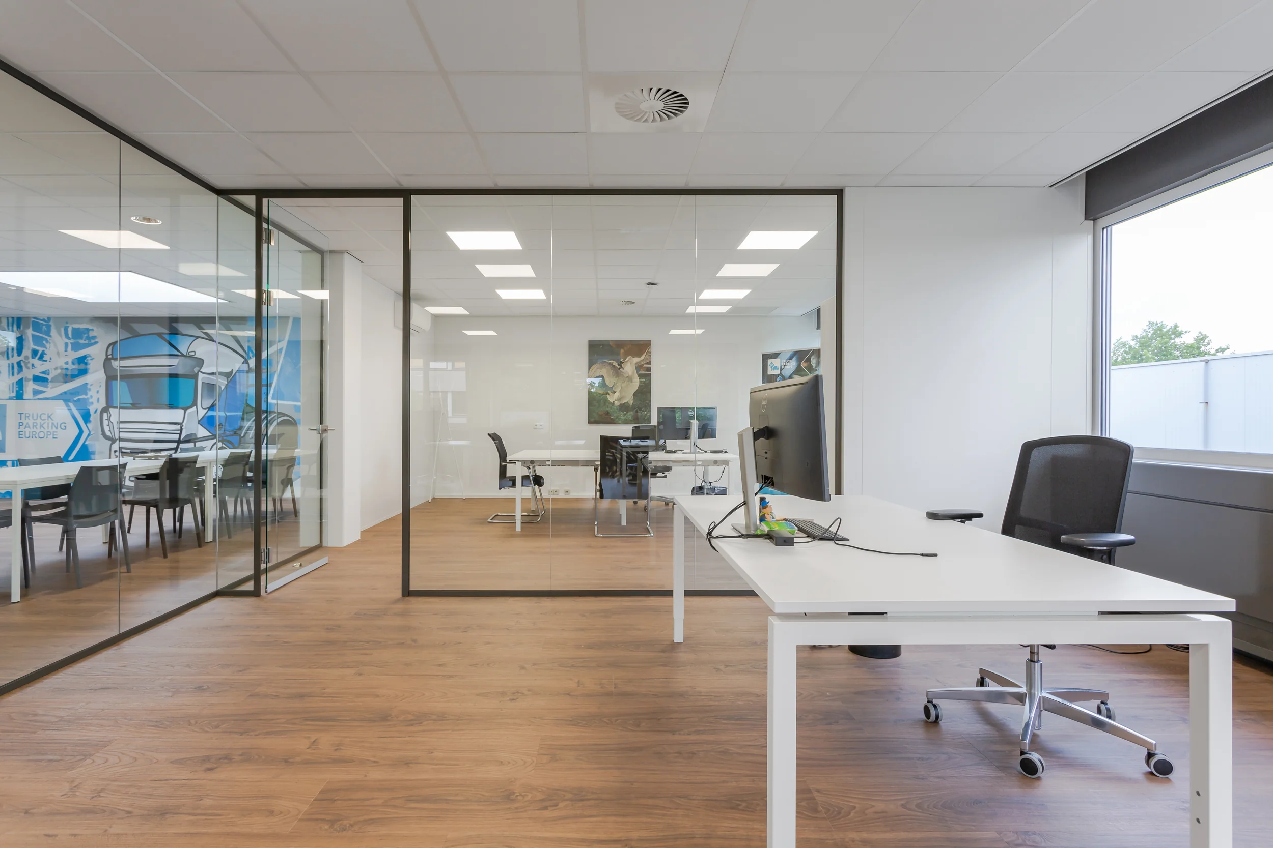 Modern kantoorinterieur met glazen vergaderruimtes, houten vloer en werkplekken met bureaustoelen.