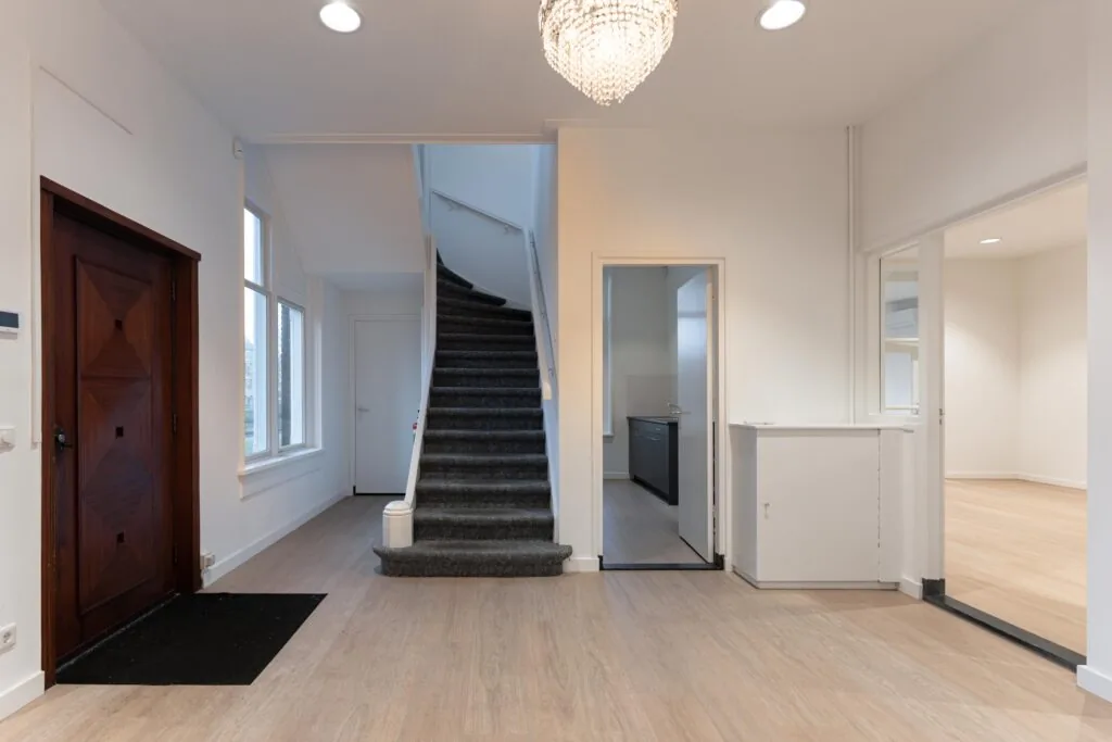 Hal met houten voordeur, trap met grijze vloerbedekking, en doorgangen naar keuken en andere kamers in een woning aan de Biltstraat.
