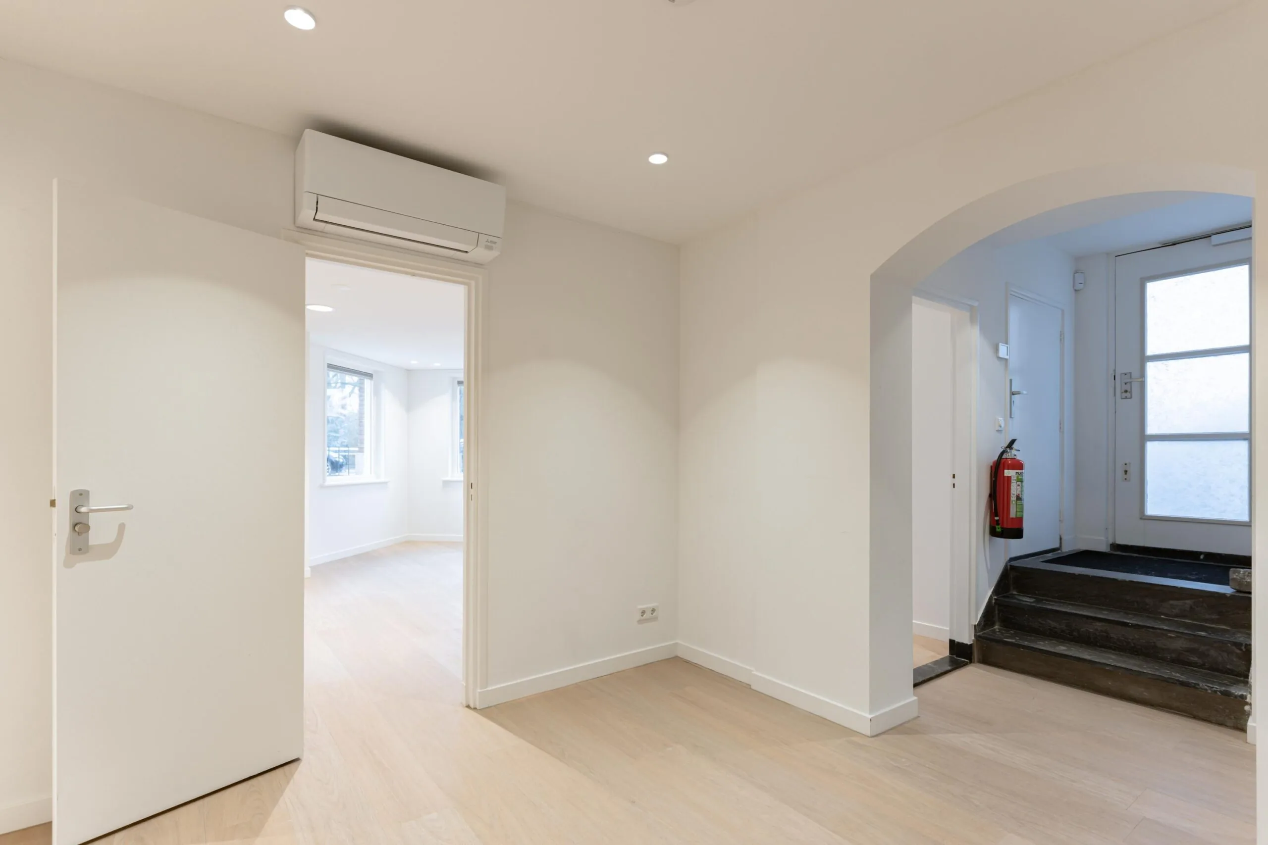 Lichte hal met een open deur naar een kamer met ramen, een airco aan het plafond en een trap bij de voordeur.