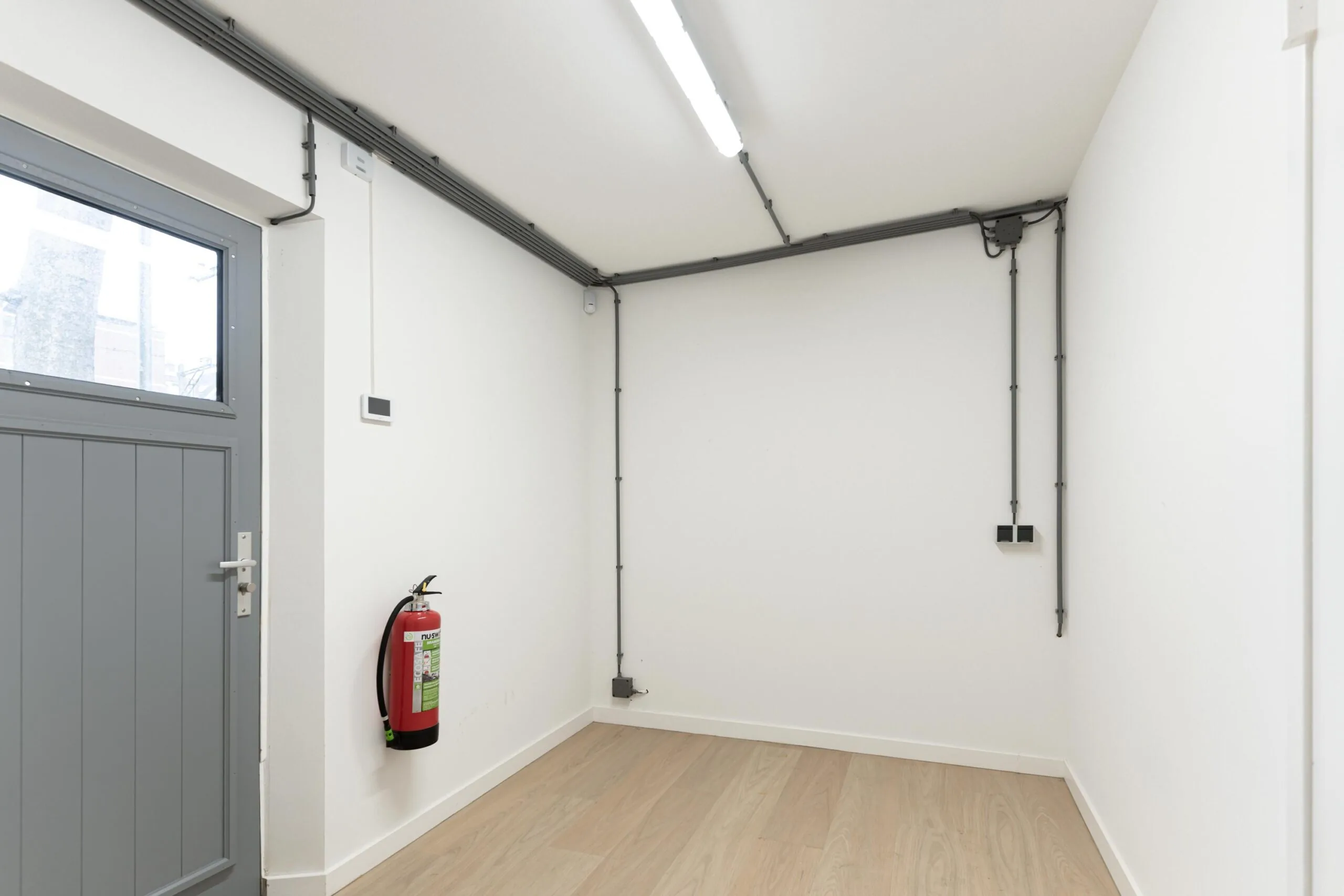Lege witte kamer met houten vloer, grijze deur, brandblusser en zichtbare elektrische bekabeling aan het plafond.