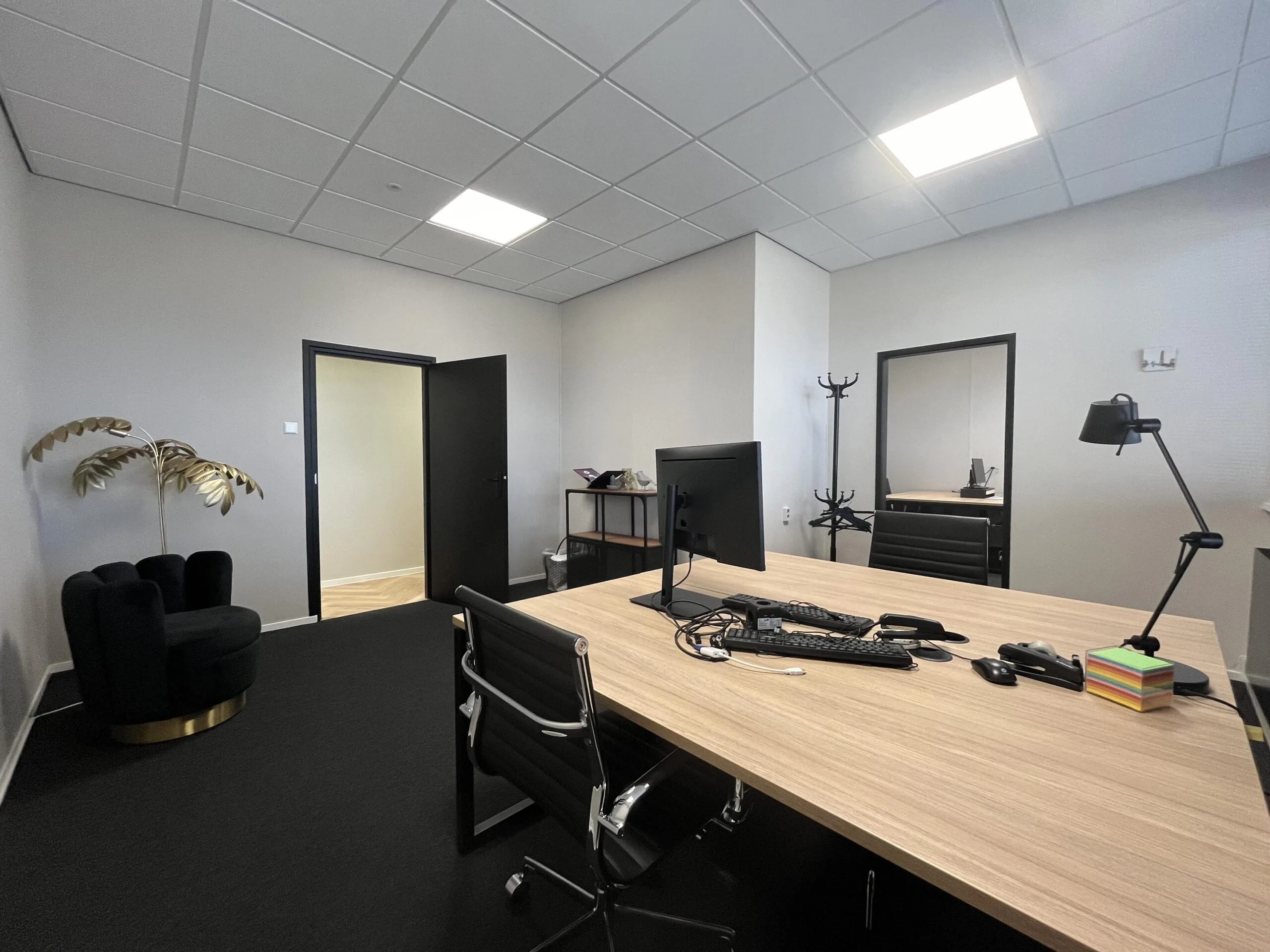Modern kantoorinterieur aan de Industrieweg met een houten bureau, bureaustoelen, computerapparatuur en een zwarte fauteuil naast een gouden decoratieve plant.