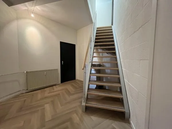 Moderne hal met een open houten trap, een zwarte deur en visgraatvloer.