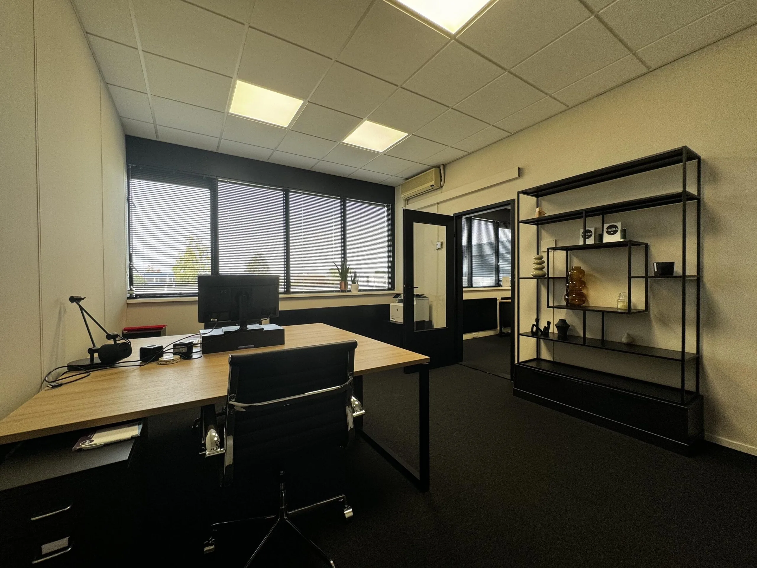 Modern ingericht kantoor met bureau, computer, open stellingkast en grote ramen met jaloezieën aan de Industrieweg.