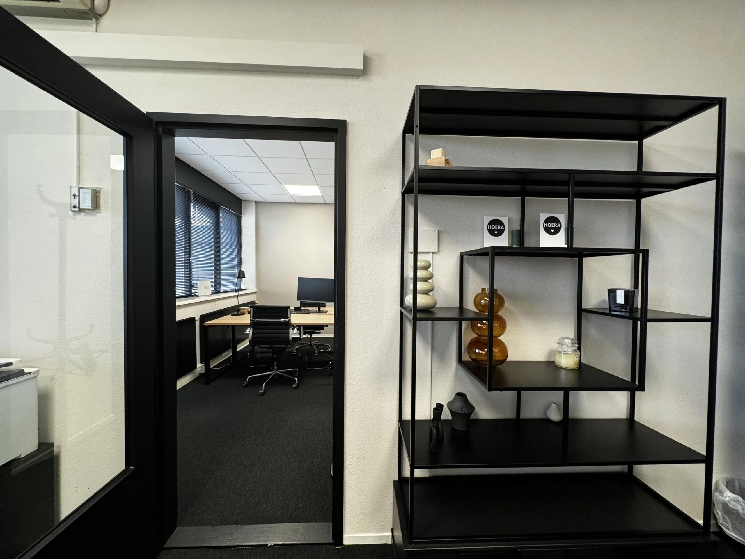 Zicht op een modern kantoor met een open deur, een zwart metalen wandrek met decoratie en een bureau met computerscherm op de achtergrond.