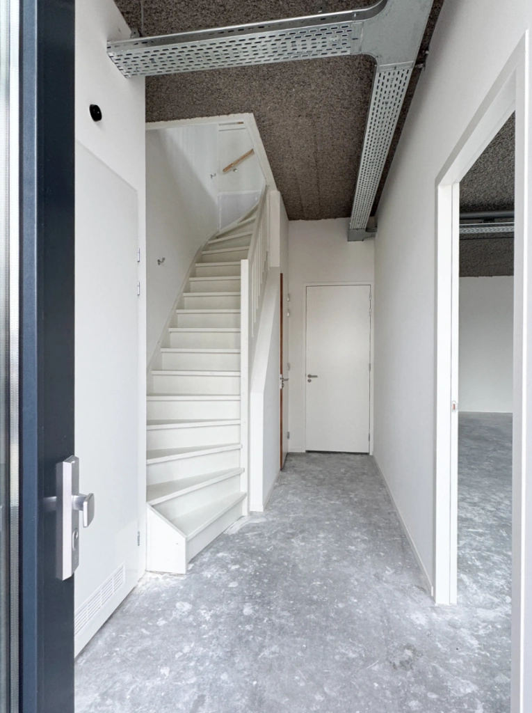 Hal met witte trap, grijze gietvloer en zicht op een naastgelegen kamer in een nieuwbouwpand aan de Nijverheidslaan.