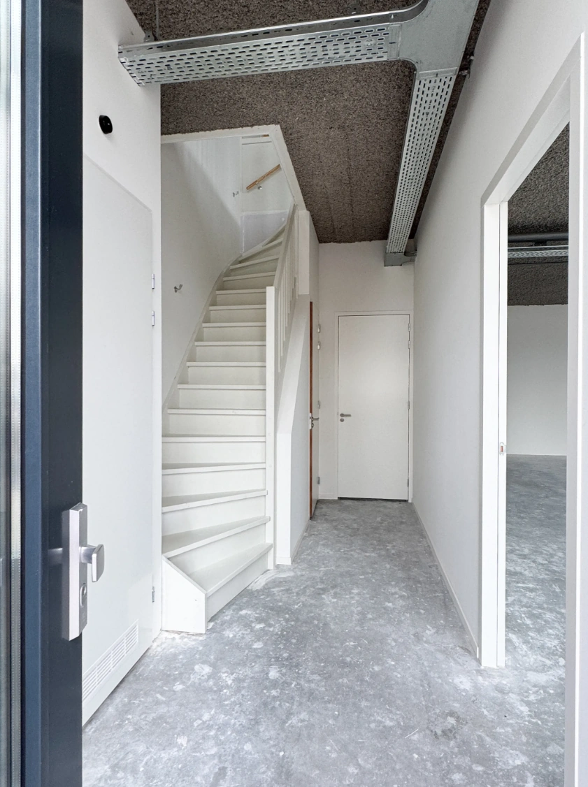 Hal met witte trap, grijze gietvloer en zicht op een naastgelegen kamer in een nieuwbouwpand aan de Nijverheidslaan.