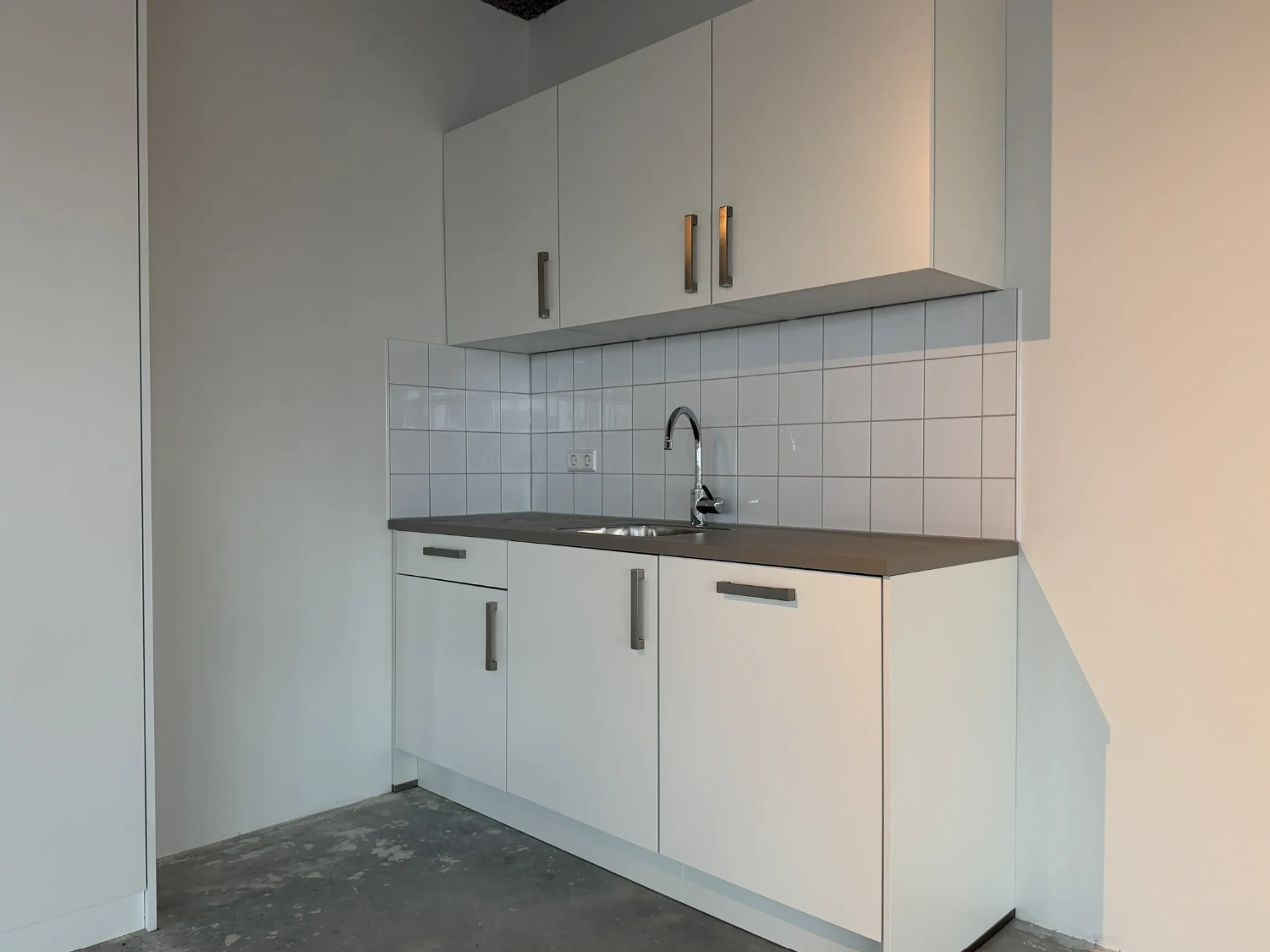 Kleine keuken met witte kastjes, grijs aanrechtblad en een betegelde achterwand in een ruimte aan de Nijverheidslaan.