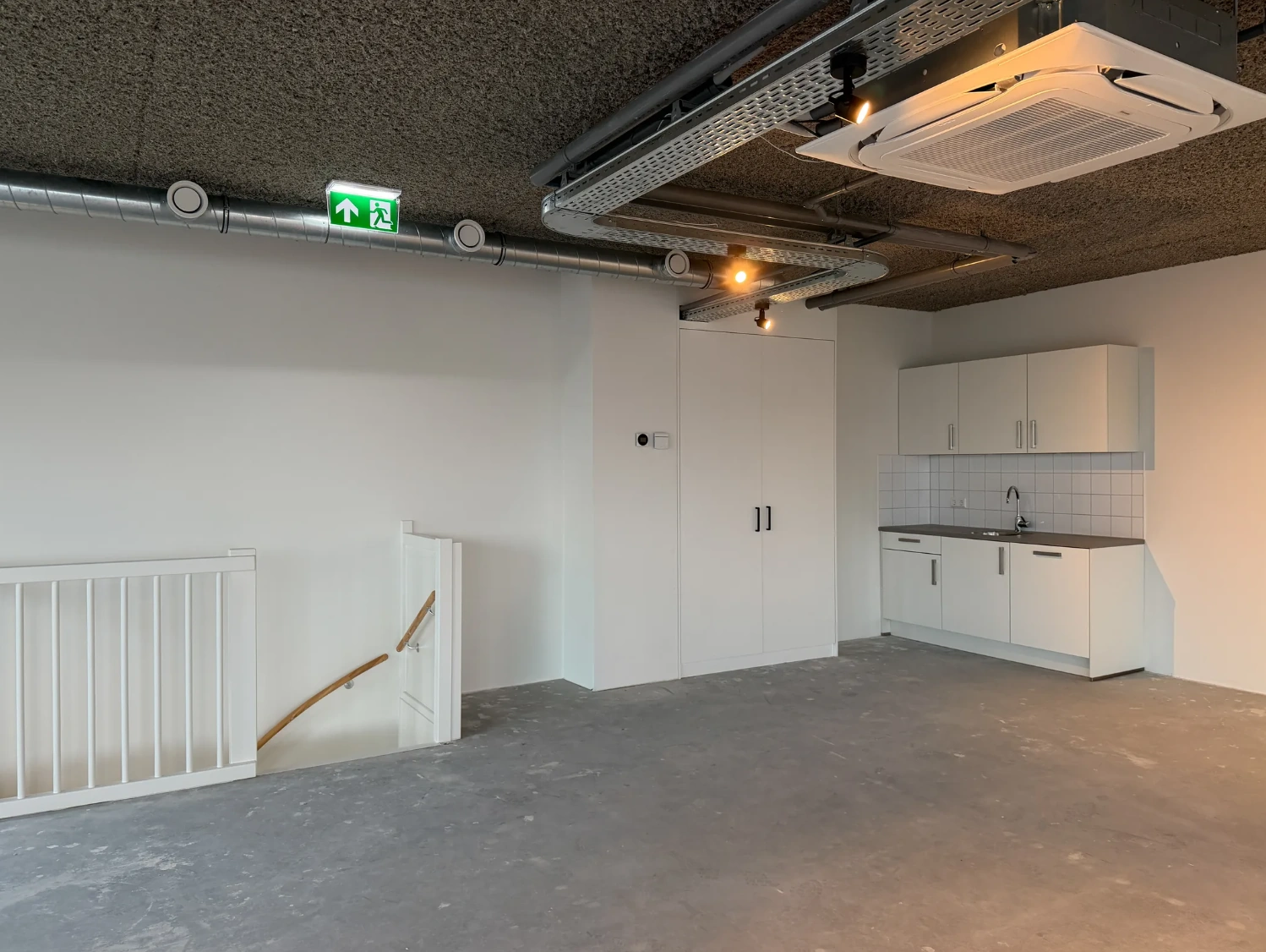 Moderne, lege kantoorruimte met pantry, trap en zichtbare ventilatiebuizen aan het plafond.