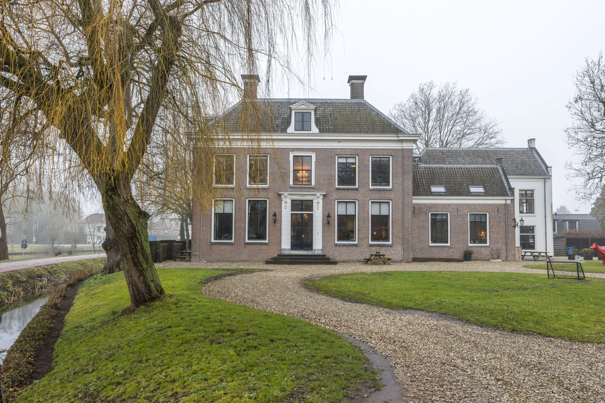 Groot statig herenhuis aan een bochtig grindpad met een sloot en wilgenboom op de voorgrond.