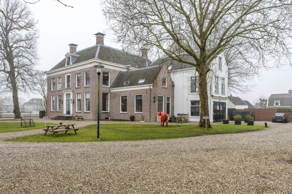 Monumentaal herenhuis aan een grindpad met picknicktafels en een rode koeiensculptuur in de tuin.