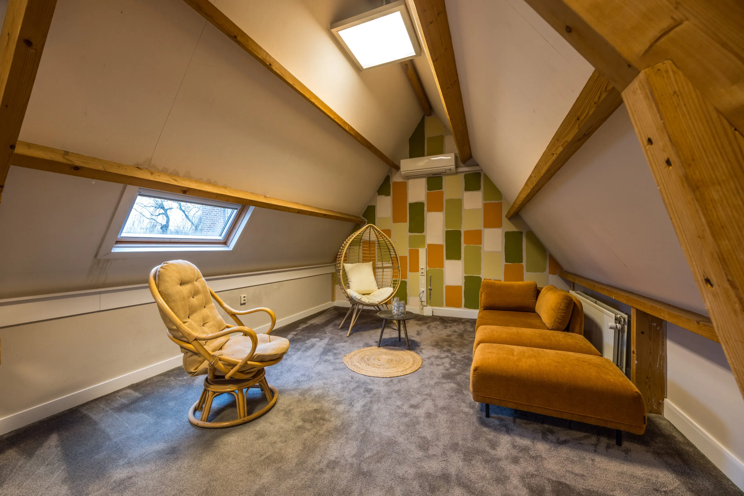 Zolderkamer met schuin dak, houten balken, een oranje chaise longue, rotan hangstoel en kleurrijke accentmuur.