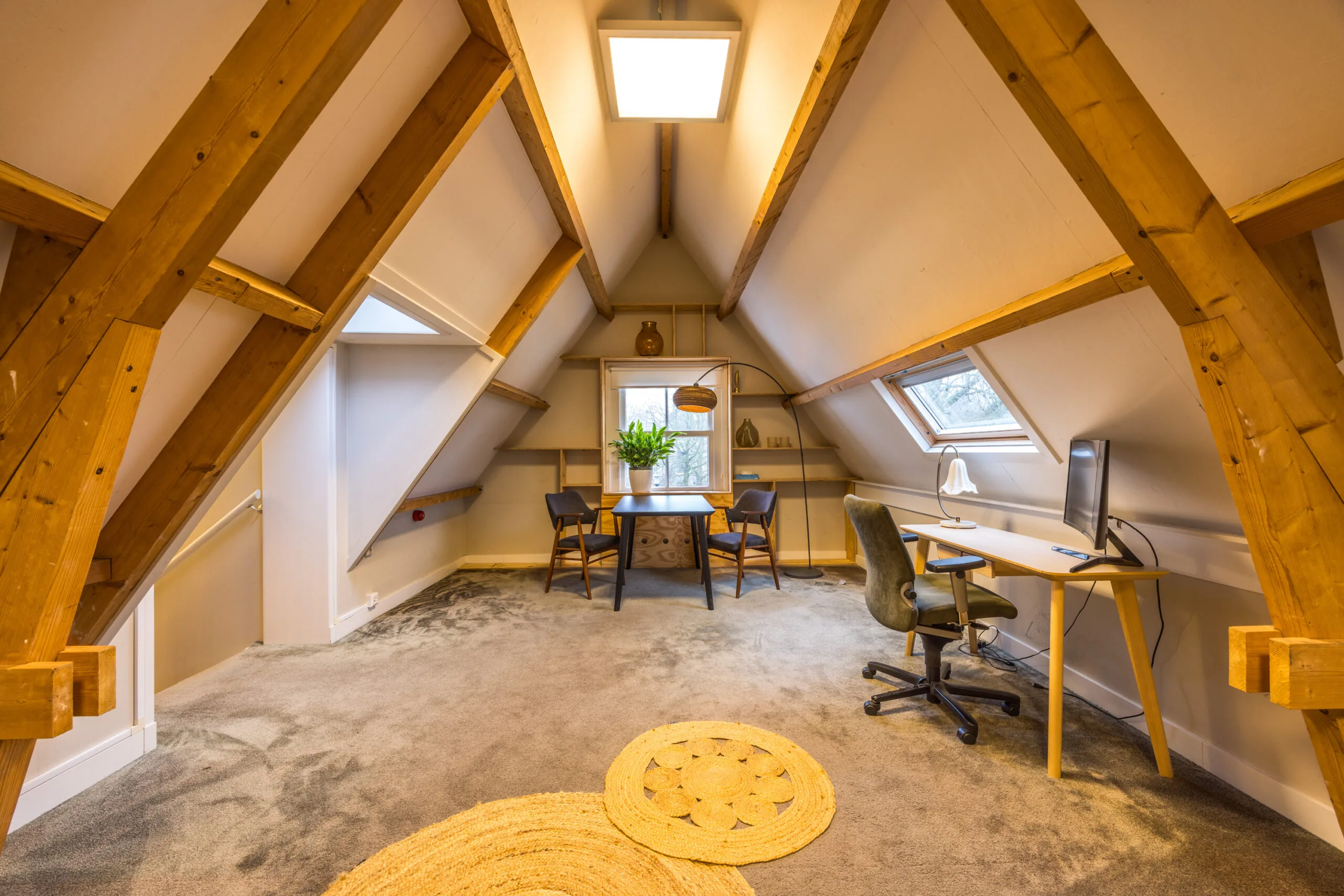Gezellige zolderkamer met houten balken, een bureau bij het raam en een zithoek met tafel en twee stoelen.